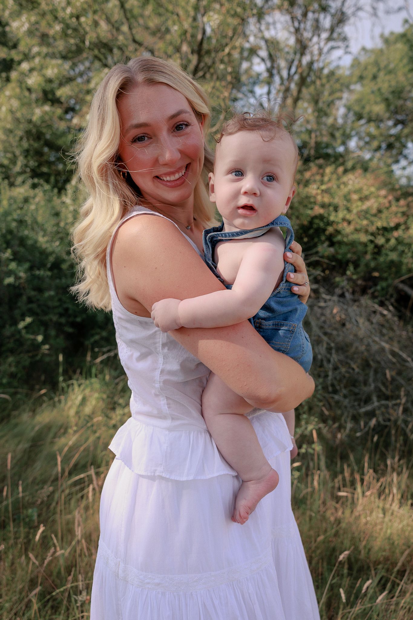 Smiling blonde woman in white dress holding a baby in denim overalls outdoors in a lush green setting.
