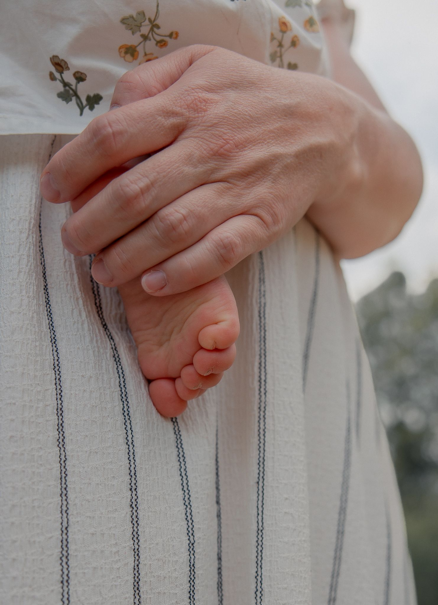 A parent gently holds a newborn baby's tiny feet against their striped linen outfit outdoors.