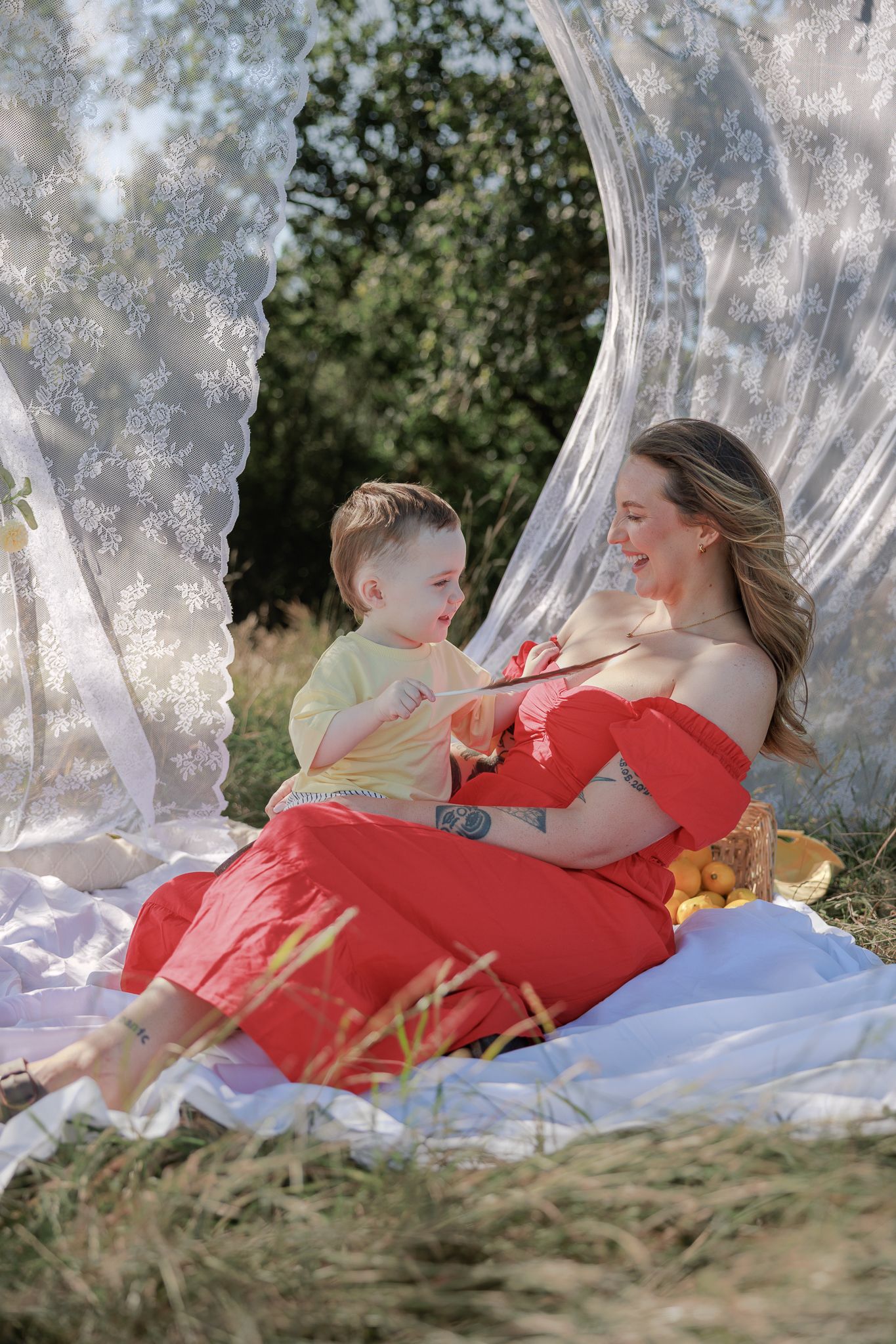 Pregnant woman in red dress sits on blanket with toddler boy, surrounded by white lace curtains outdoors in Fairford, Cotswolds.