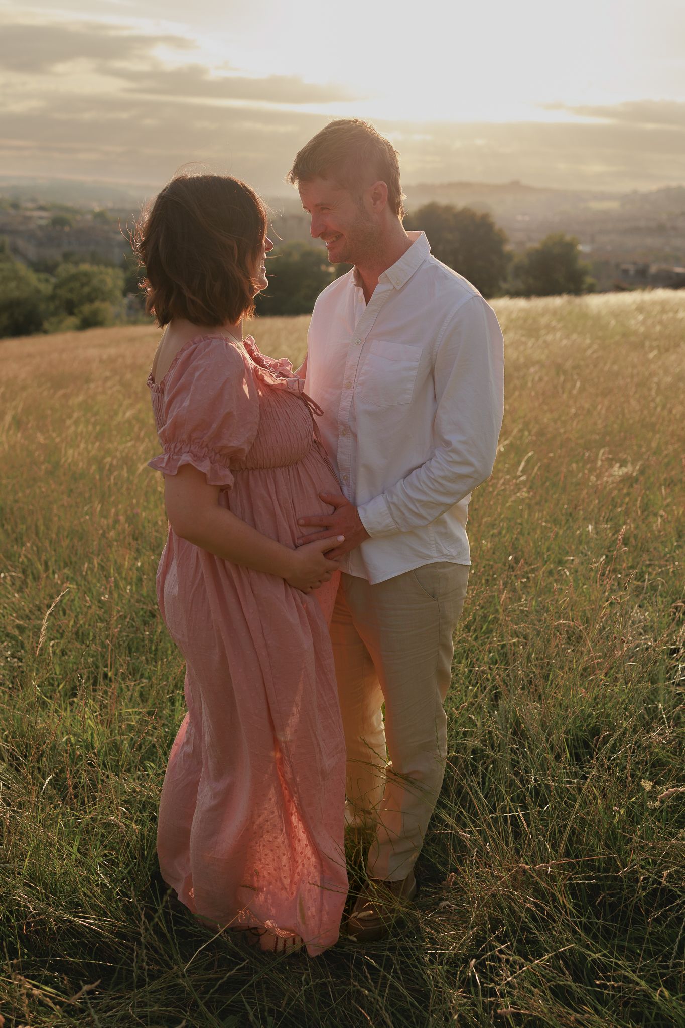 Couple sharing a tender moment in a golden sunset field, woman in pink maternity dress cradling baby bump for a Maternity Photoshoot, Bath.