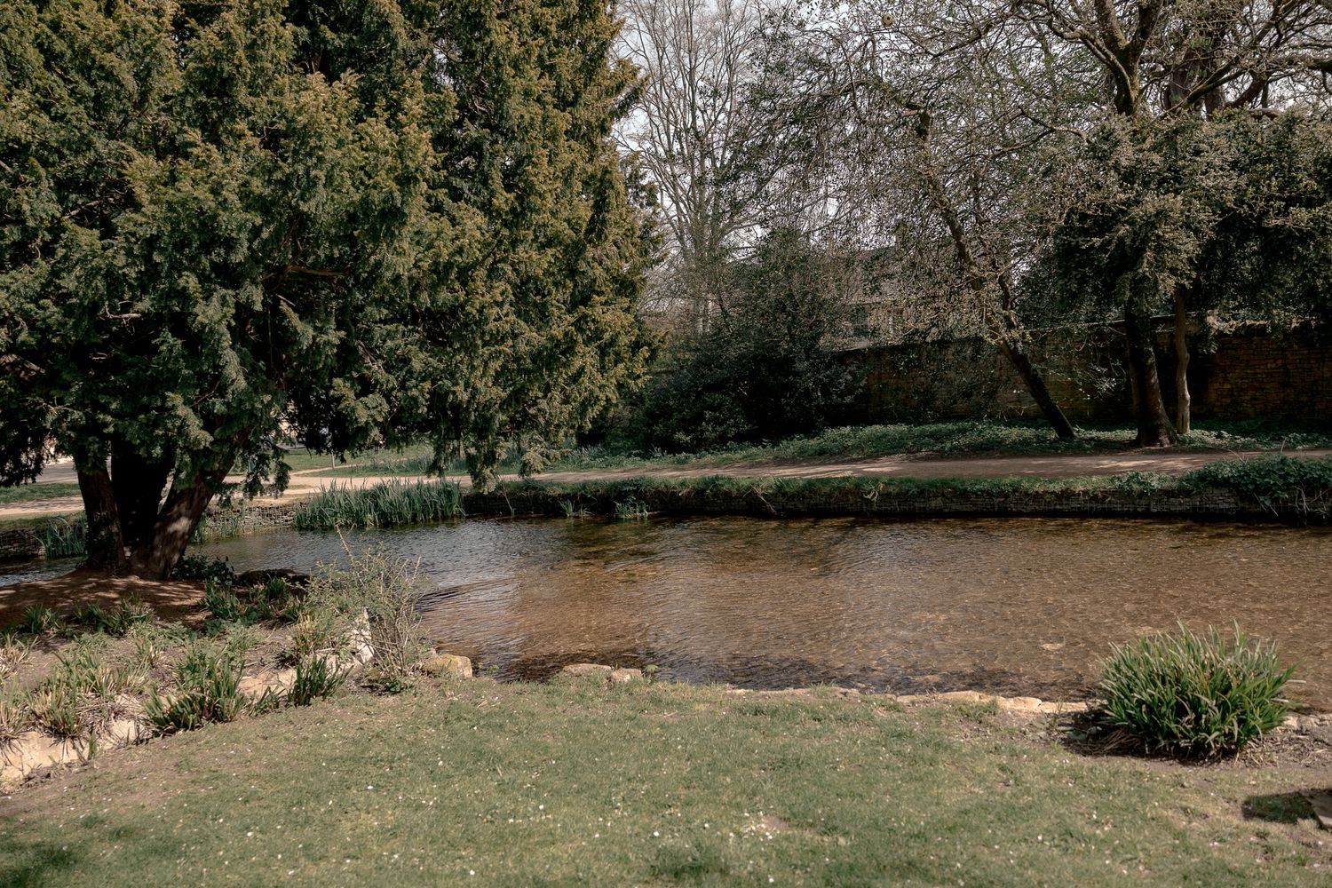 Serene garden pond surrounded by lush green trees and grass in a tranquil outdoor setting in Lower Slaughter, Cotswolds, UK.