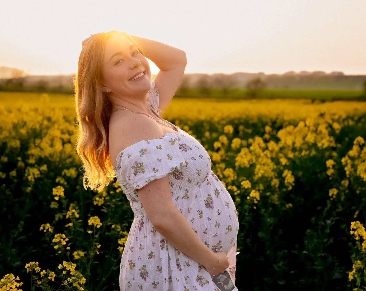 Smiling pregnant woman in white floral dress standing in yellow rapeseed field at golden sunset in Chipping Campden, Cotswolds.