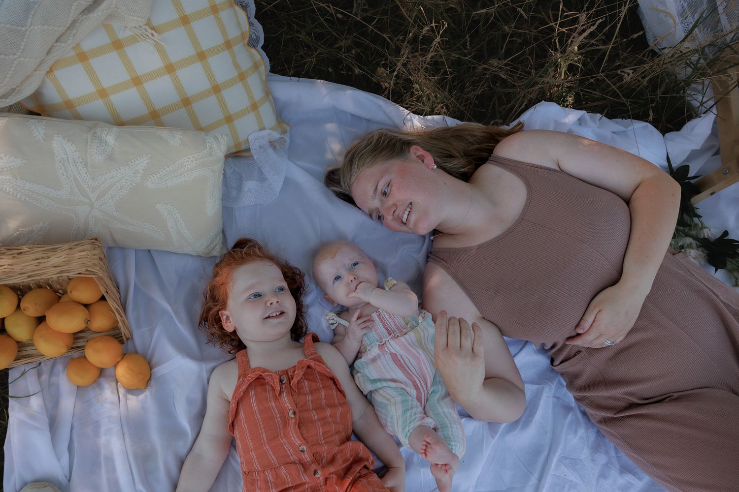 A mother and two young children lying on a white picnic blanket outdoors, with oranges and a basket nearby.