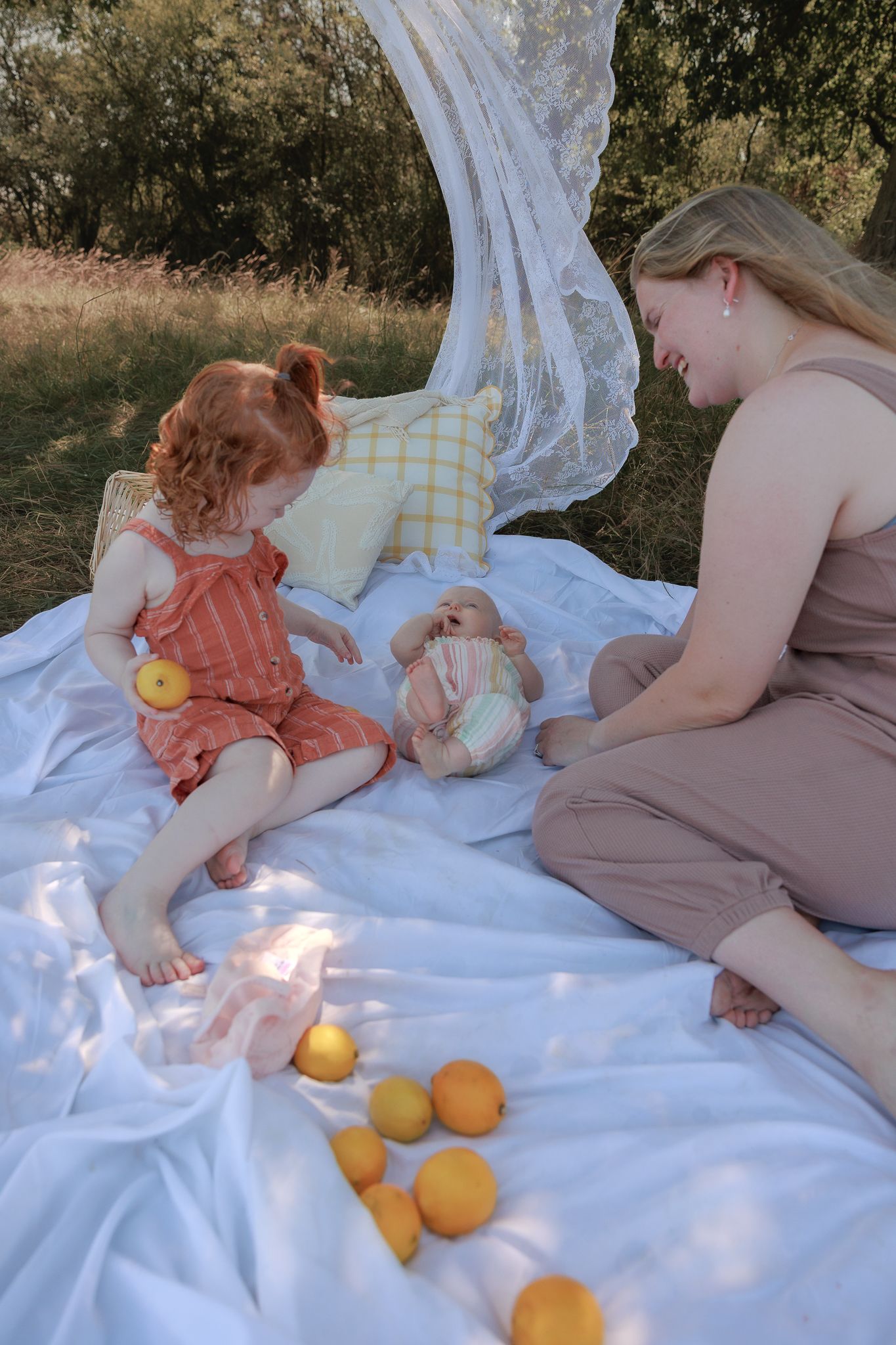 Mother and toddler girl enjoying a sunny outdoor picnic on white blanket with oranges and pillows in nature in Henley-on-Thames, Oxfordshire