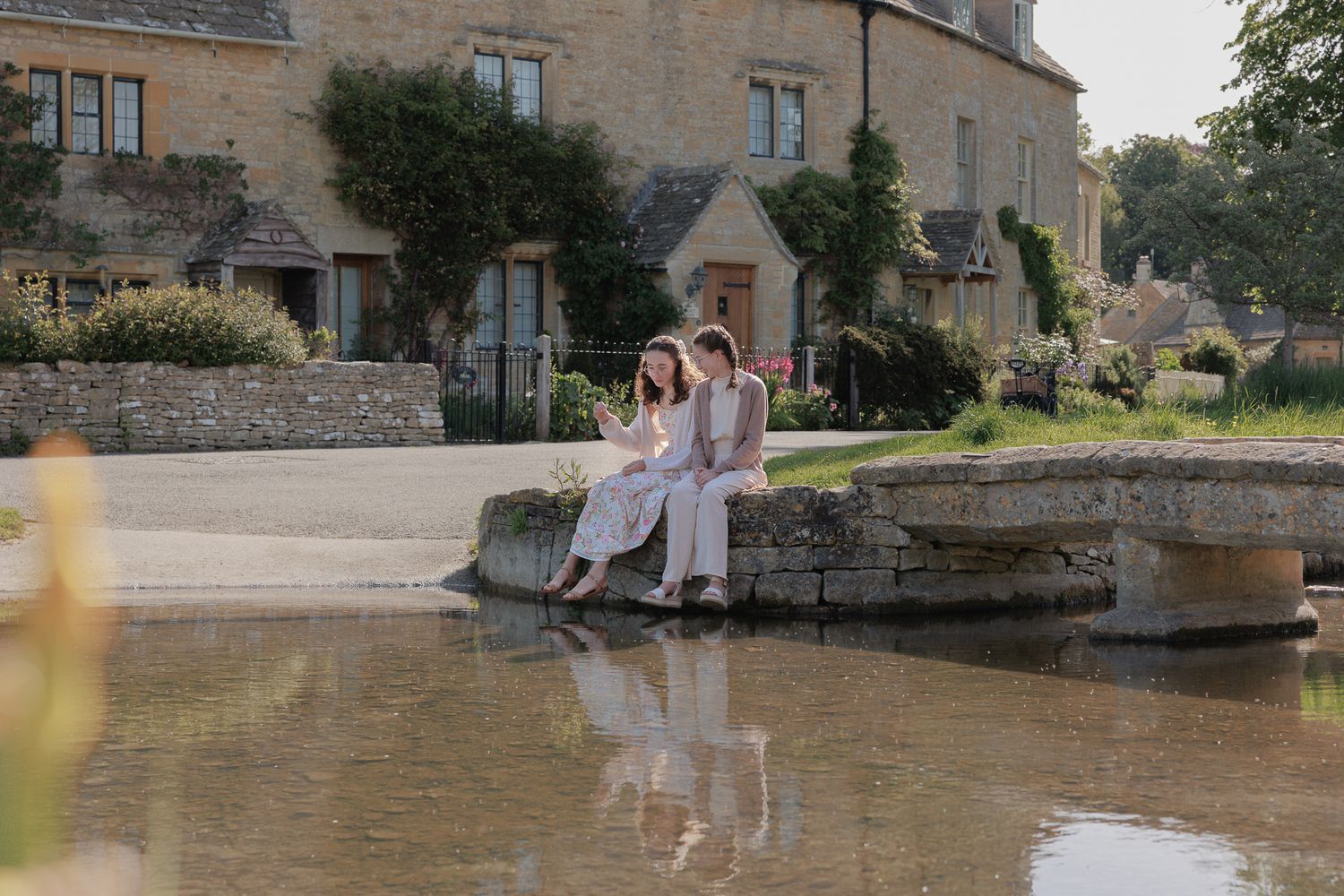 Couple sitting together on a stone bridge over a calm pond beside a charming Cotswolds village.