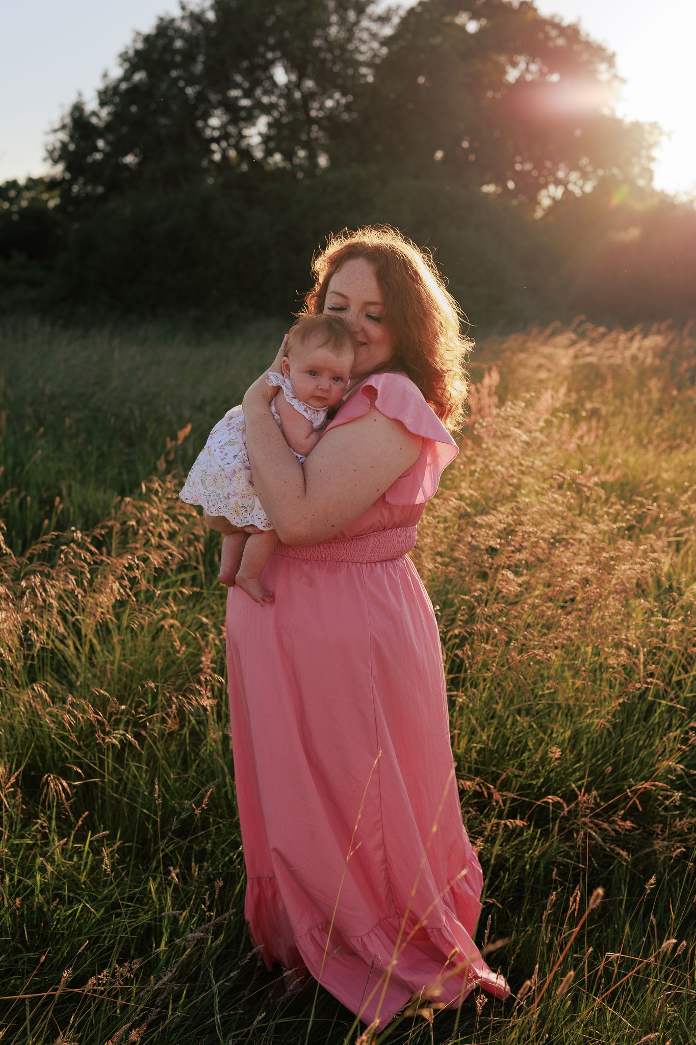 A mother in a pink maxi dress tenderly holds her baby in a sunlit field at golden hour in Hampshire.