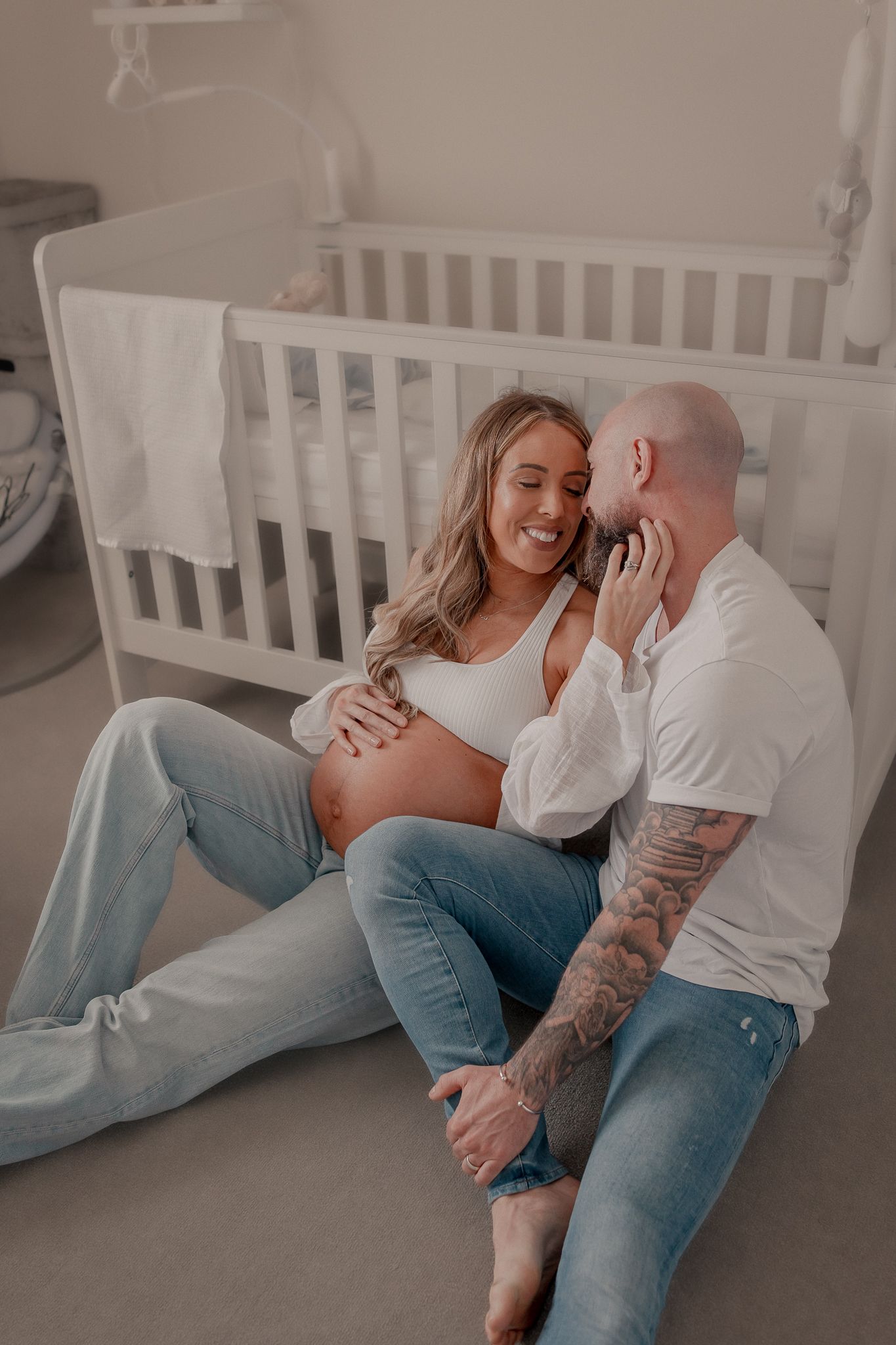 Pregnant couple sitting on nursery floor by white crib, sharing a tender moment, man with tattoos kissing woman's cheek in Quenington.