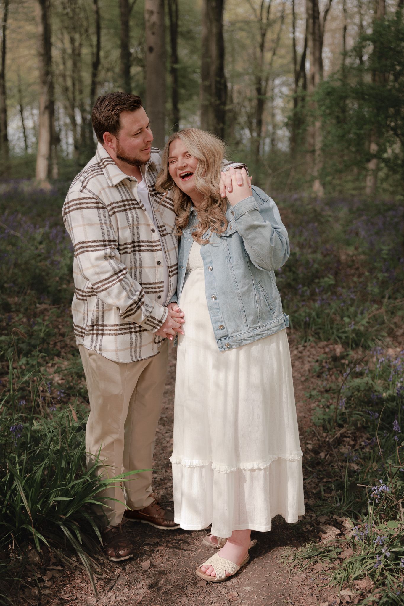 A laughing couple holds hands on a woodland path surrounded by bluebells, she wears a white dress and denim jacket.