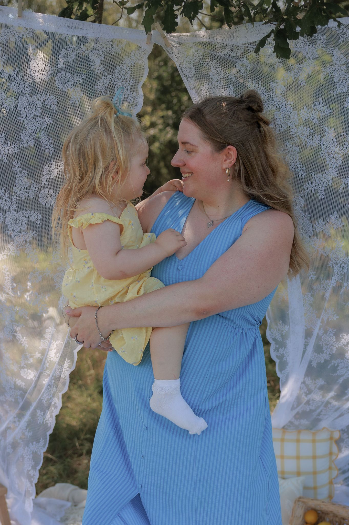 A smiling woman in a blue striped dress holds a toddler girl in a yellow dress outdoors near lace curtains in Bibury, Cotswolds.