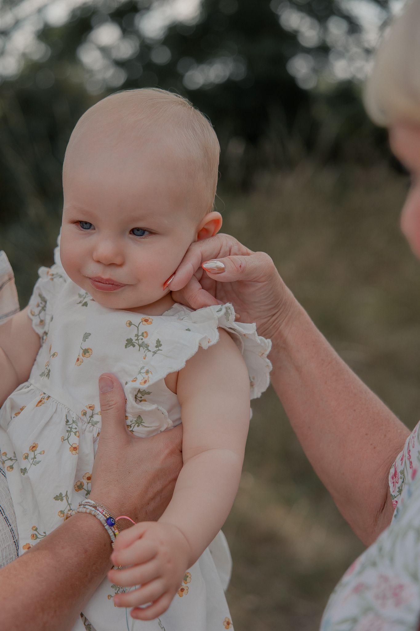 Elderly hands gently holding a smiling baby girl in a white floral dress outdoors.