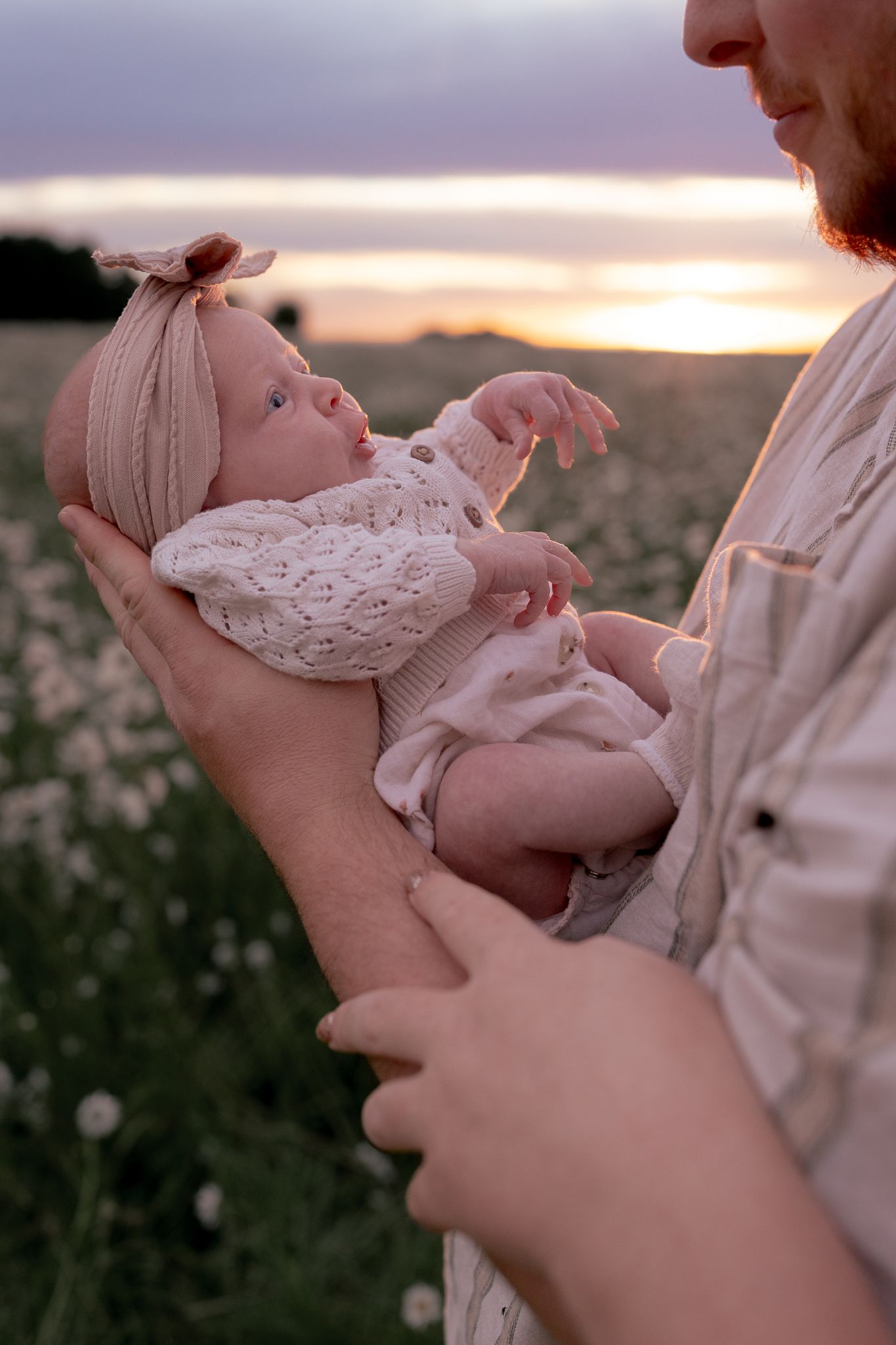 Father tenderly holding newborn baby in a sunlit daisy field at golden hour sunset.