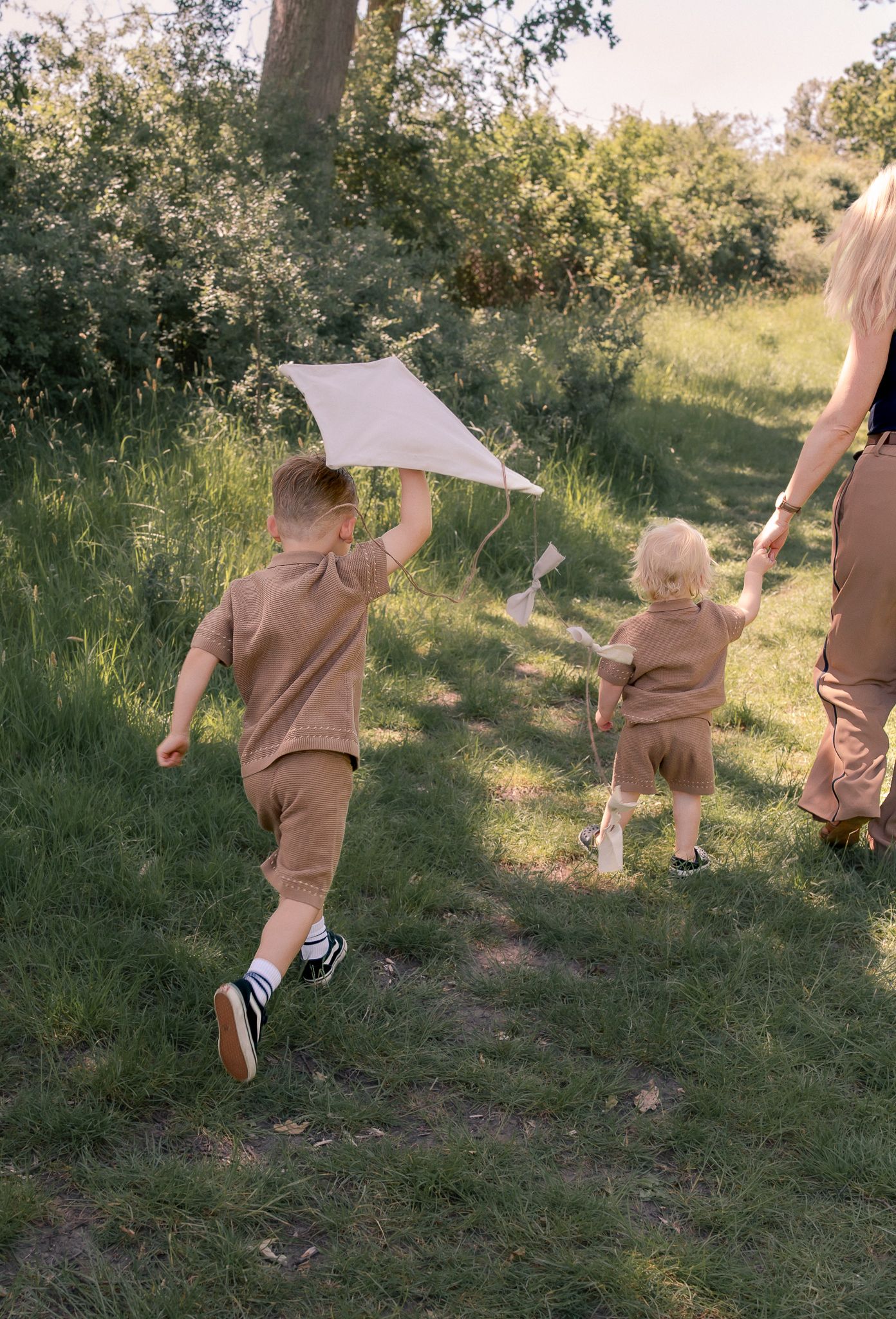 Two children running through a grassy field flying a white kite, accompanied by an adult on a sunny day.