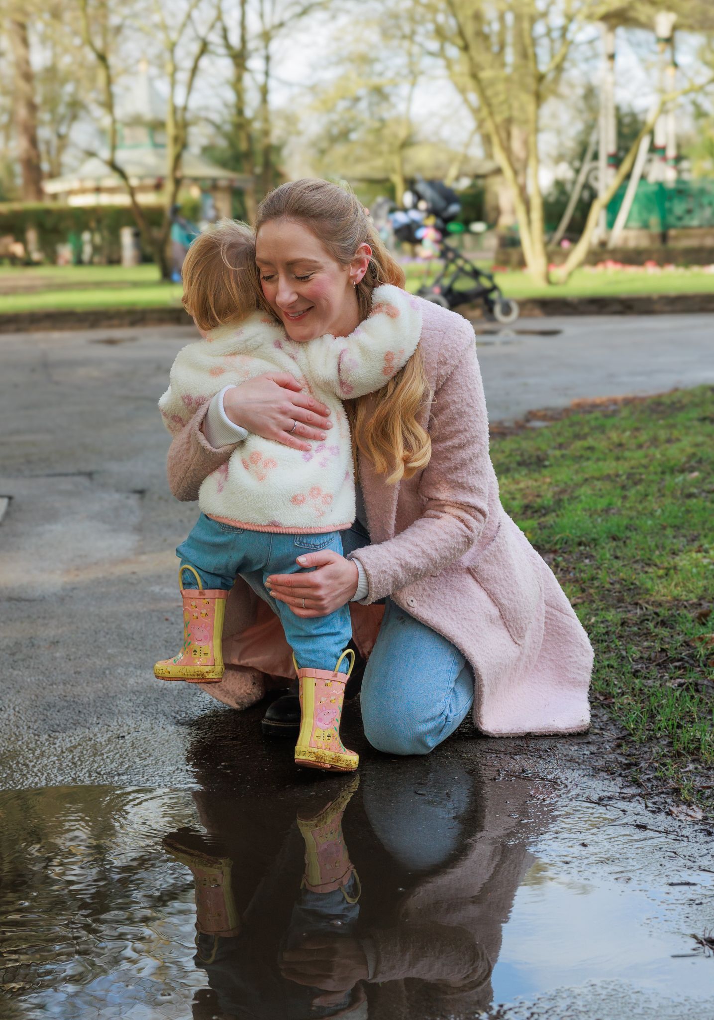 A mother crouches to hug her toddler in yellow rain boots beside a puddle in a park in North Cerney, Cotswolds.