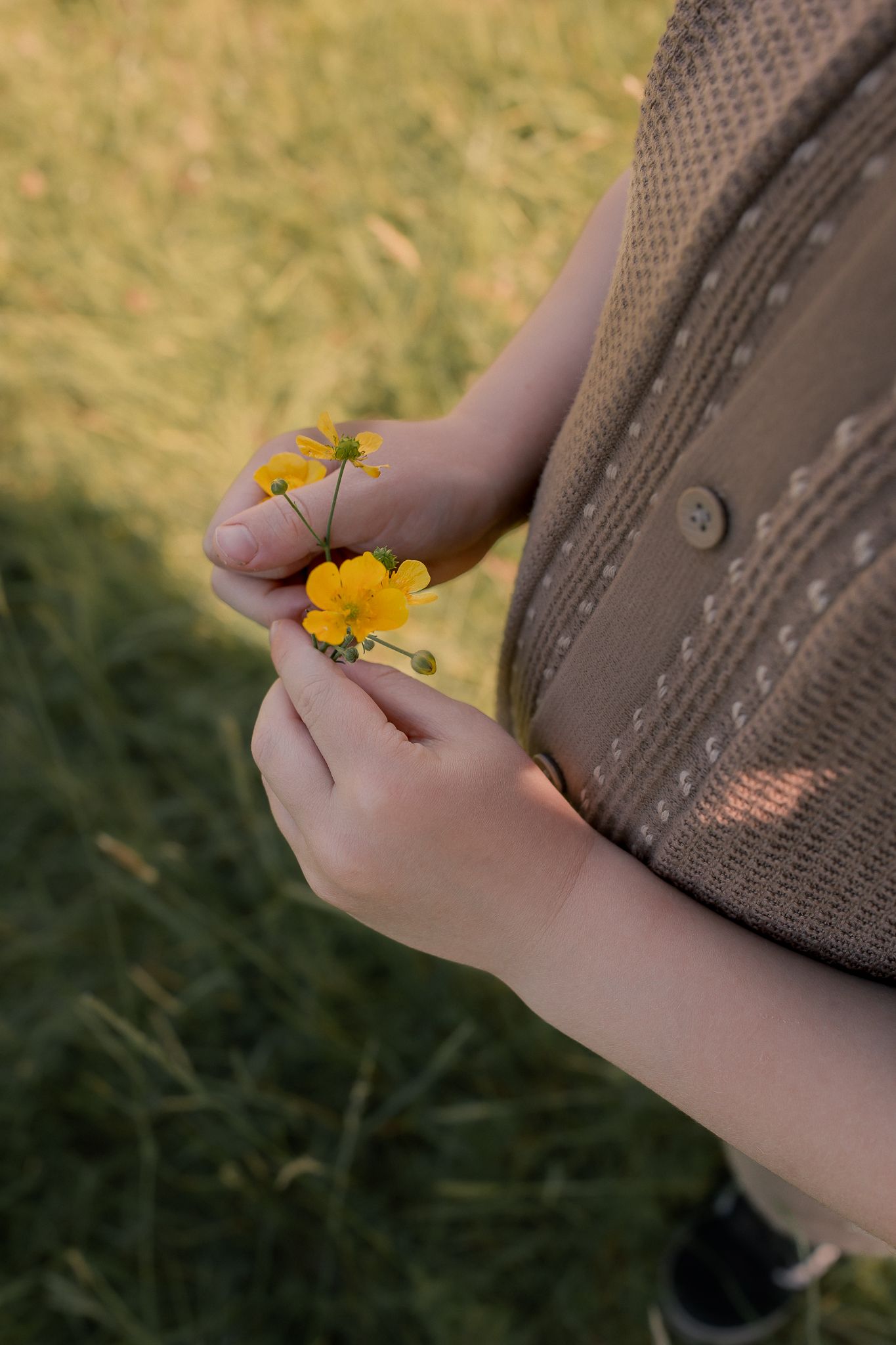 Person in beige cardigan holding small yellow wildflowers in their hands while standing in a grassy field outdoors.
