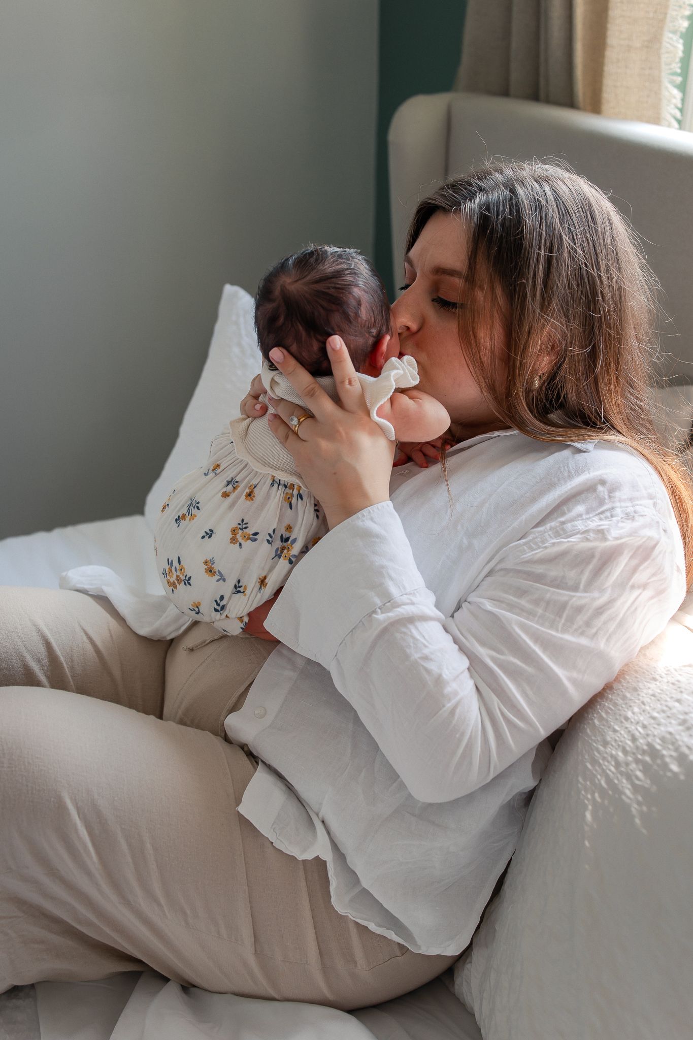 A young mother sitting in a chair gently kissing her newborn baby dressed in a floral outfit in Lacock.