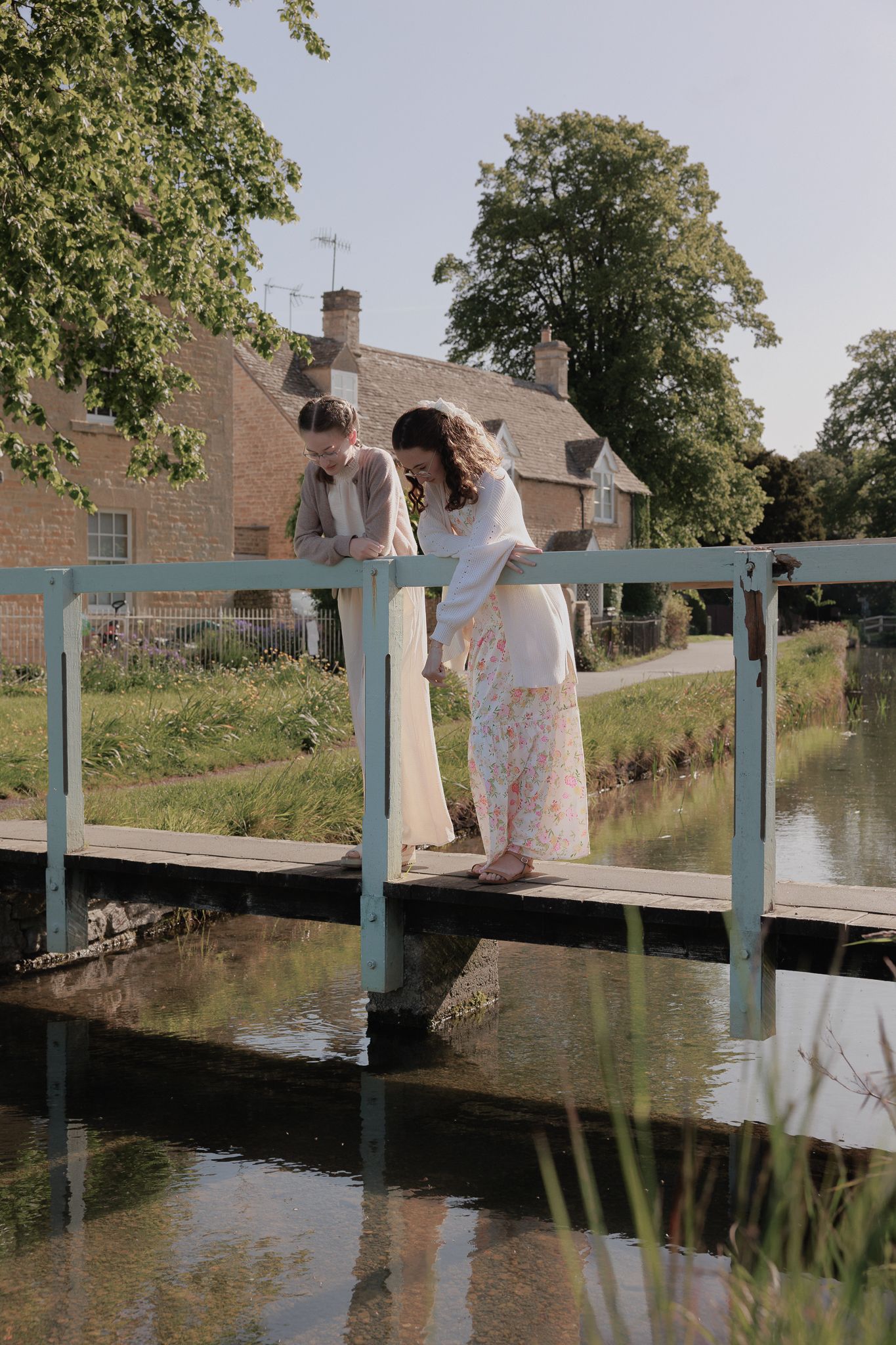 Two women in floral dresses lean over a rustic bridge railing above a calm river near a stone cottage near The Slaughters Manor House.