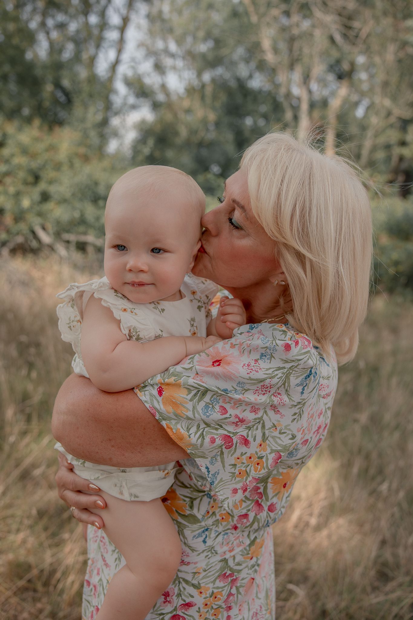 Blonde woman in floral dress kissing a baby outdoors in a natural grassy setting.