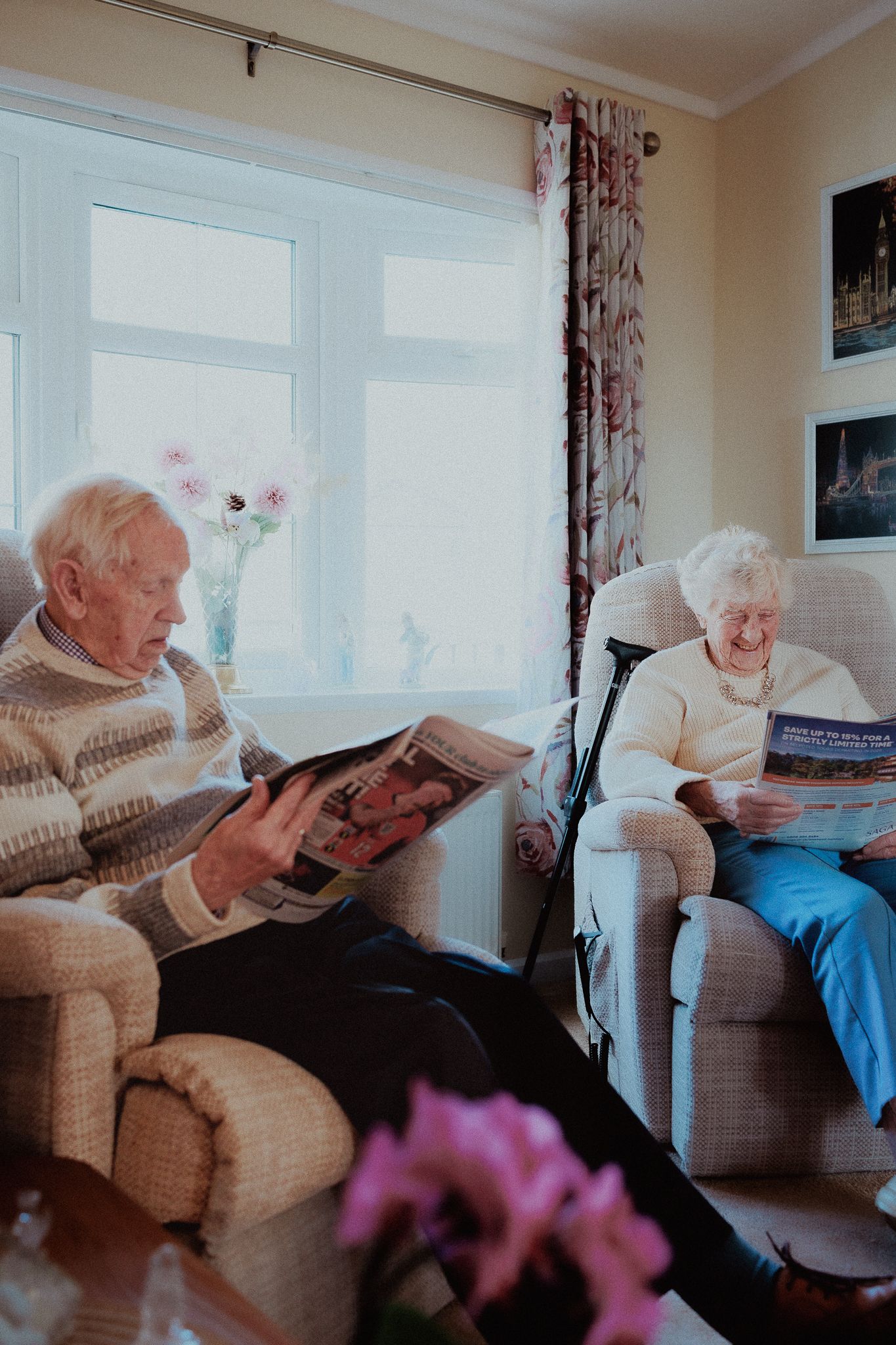 Two elderly people relaxing in armchairs reading newspapers in a cosy living room in Lower Slaughter.