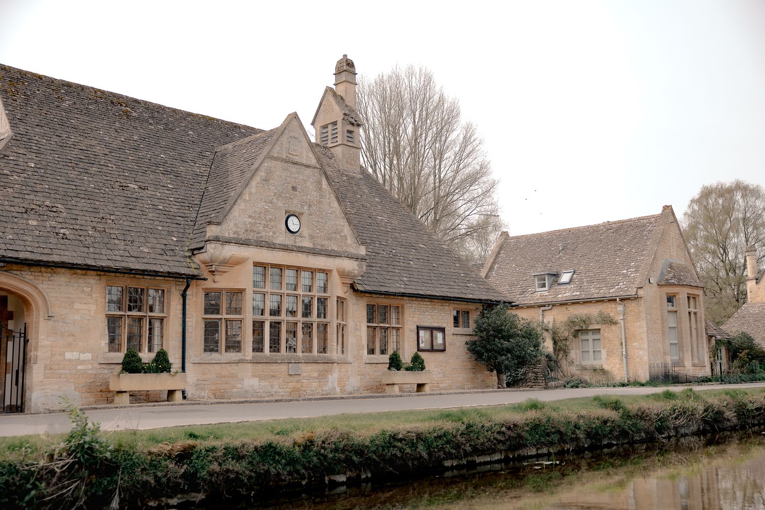 Historic Cotswold stone building with large windows beside a calm river, surrounded by bare winter trees in Lower Slaughter, Cotswolds, UK.