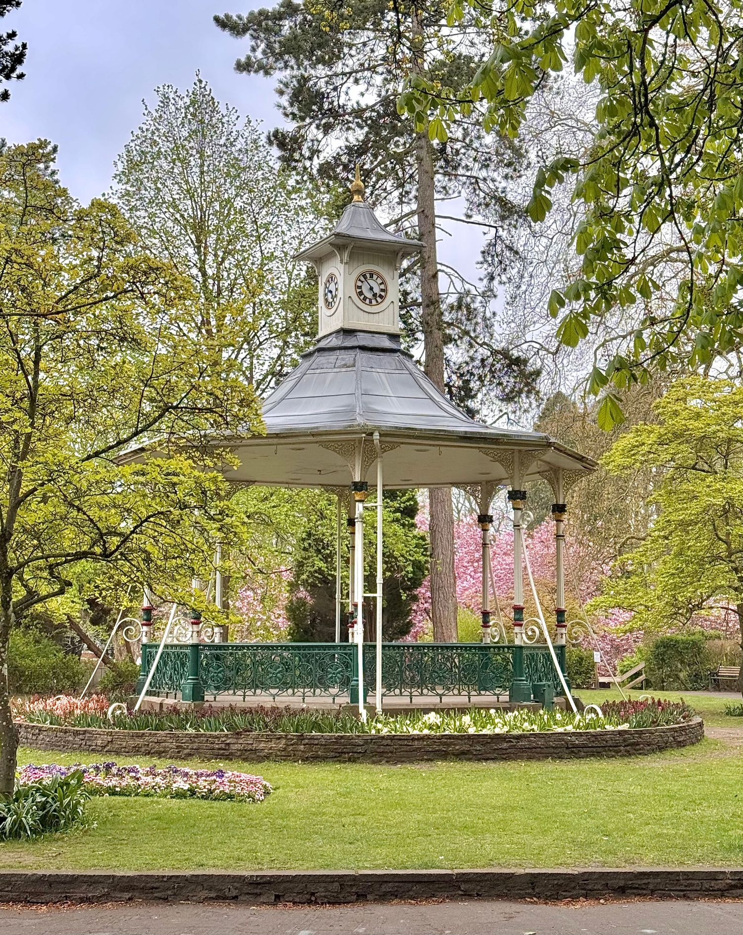 Victorian bandstand with clock tower surrounded by colorful spring gardens and lush green trees in a public park in Old Town Gardens.