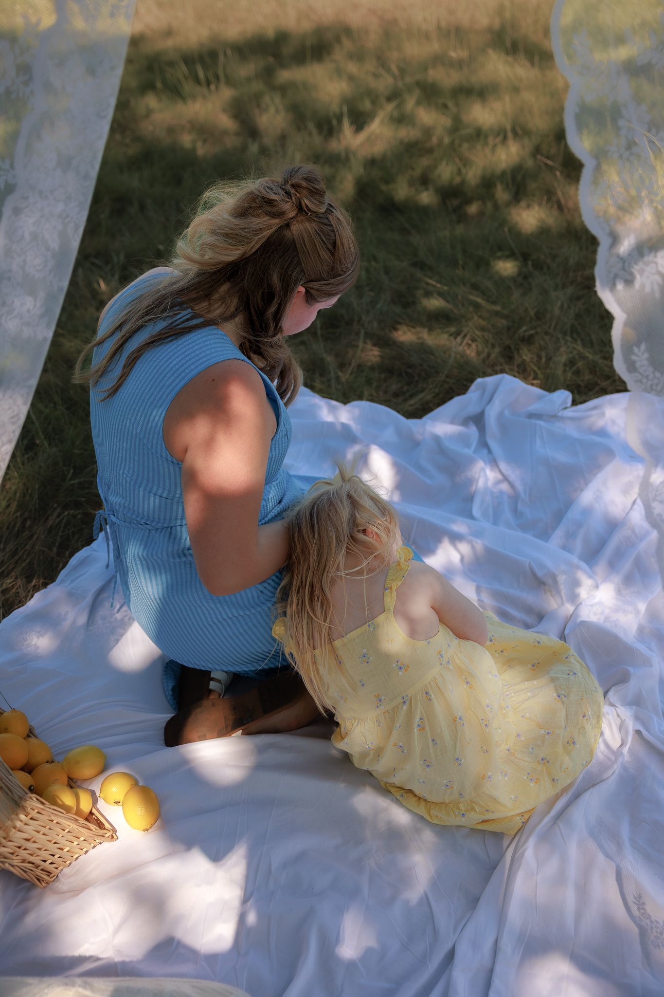 Mother and daughter in blue and yellow dresses sitting on white blanket outdoors with basket of lemons in Bibury, Cotswolds.