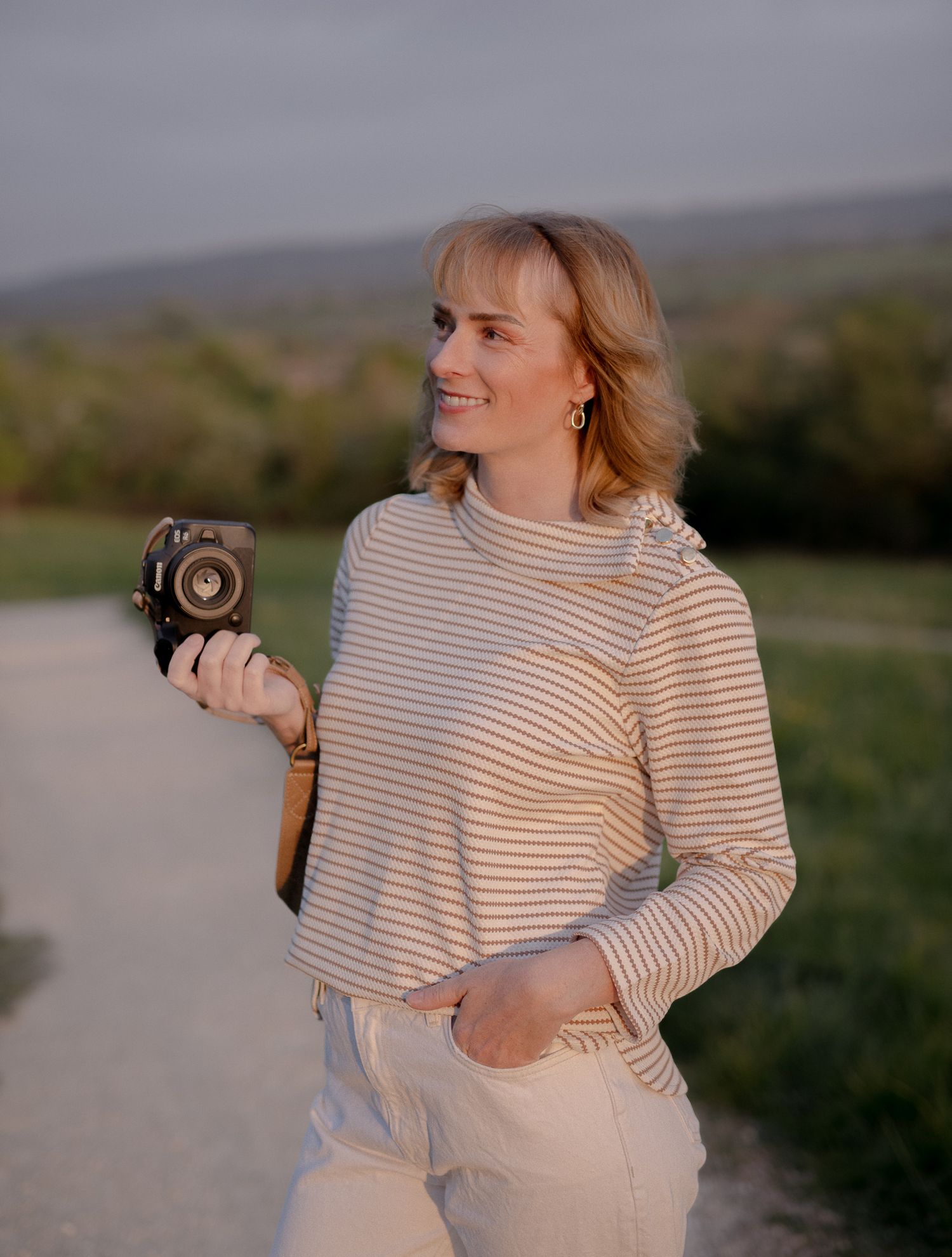 Smiling woman holding a vintage camera outdoors on a path, wearing a striped shirt in warm golden hour light.