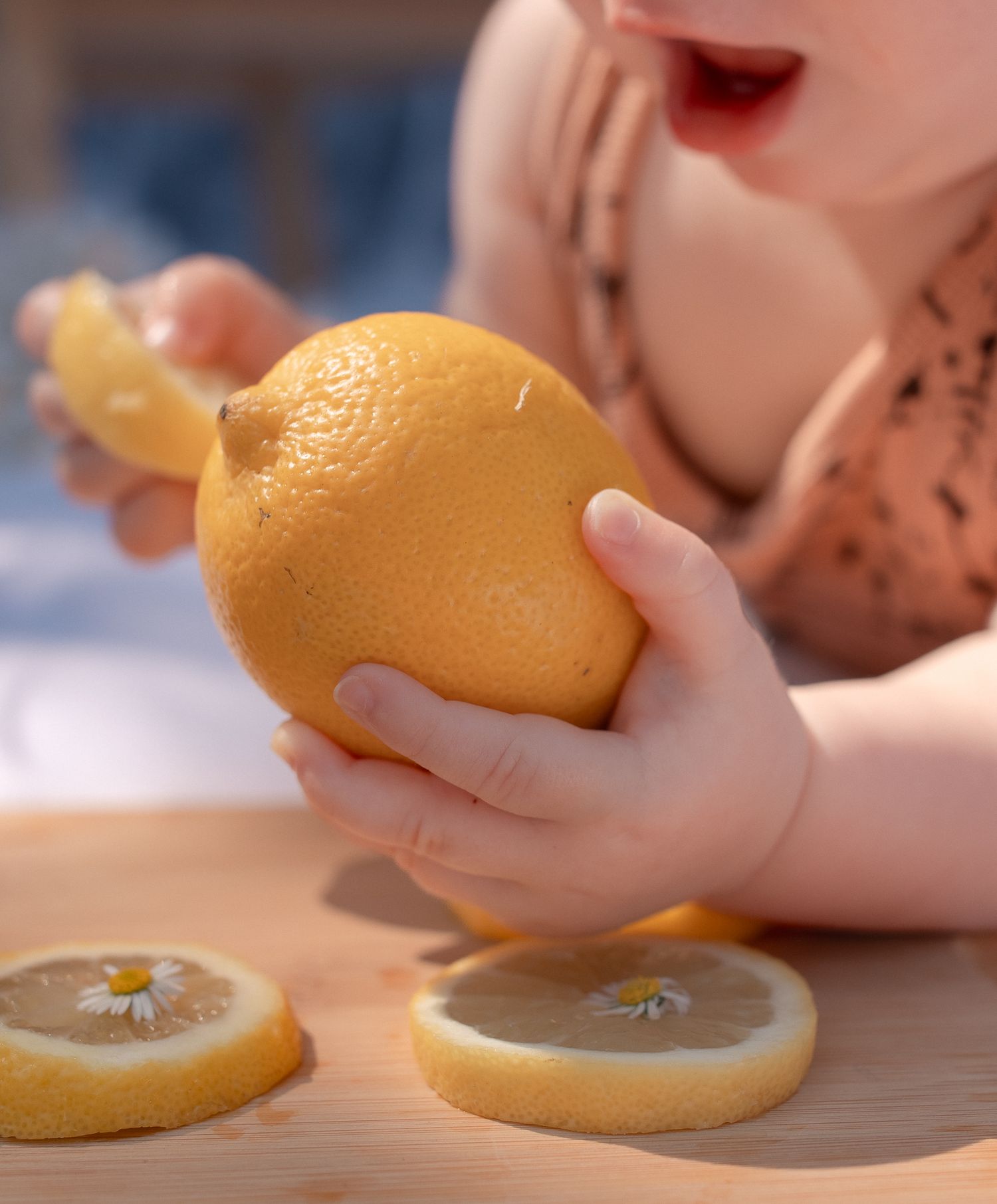 A baby holds a large lemon with sliced lemon pieces on a wooden table, mouth open in curiosity in Burford, Cotswolds.