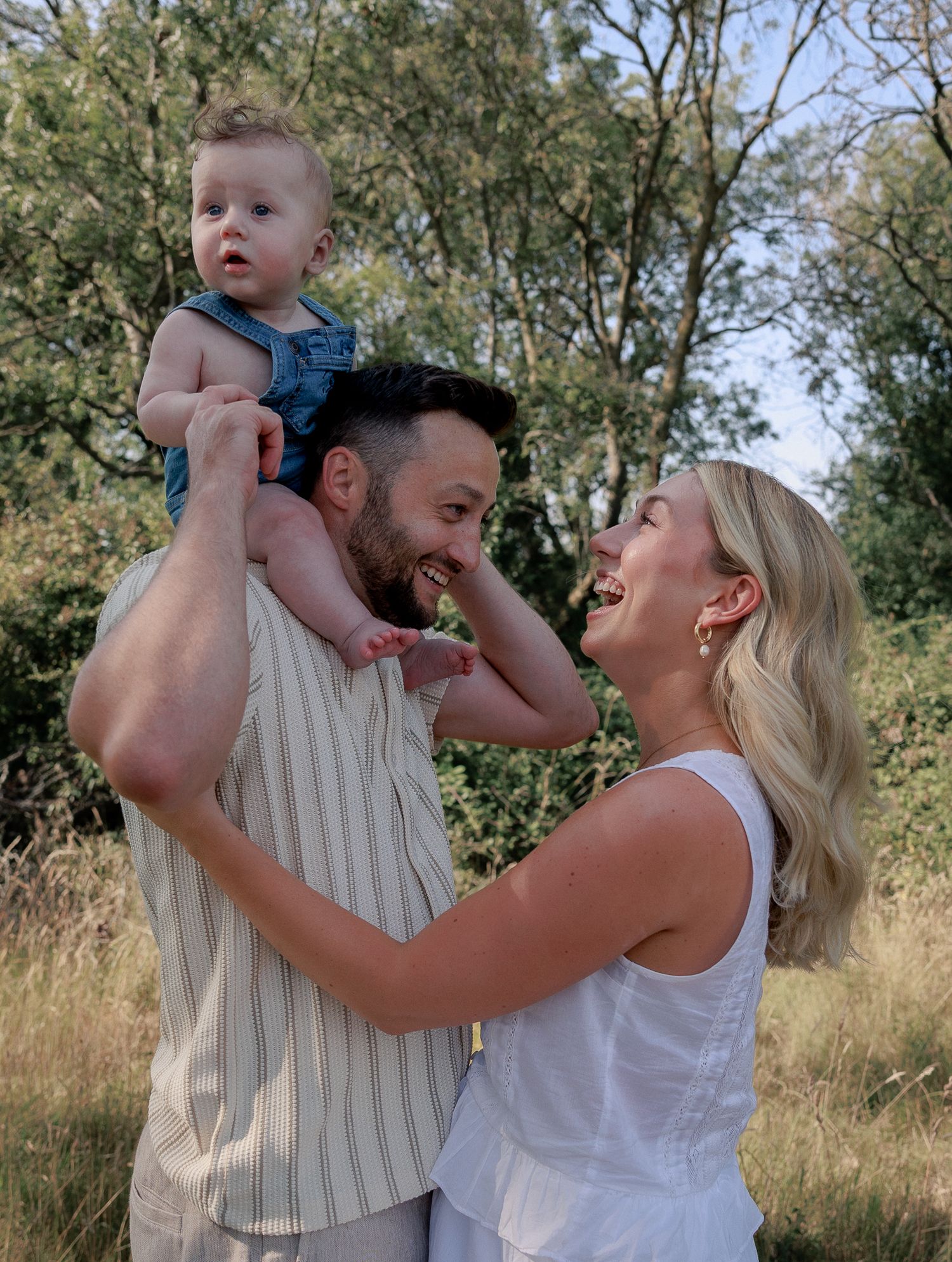 Happy family with man lifting baby while smiling woman in white dress enjoys outdoor moment in sunny field in Burford, Cotswolds.