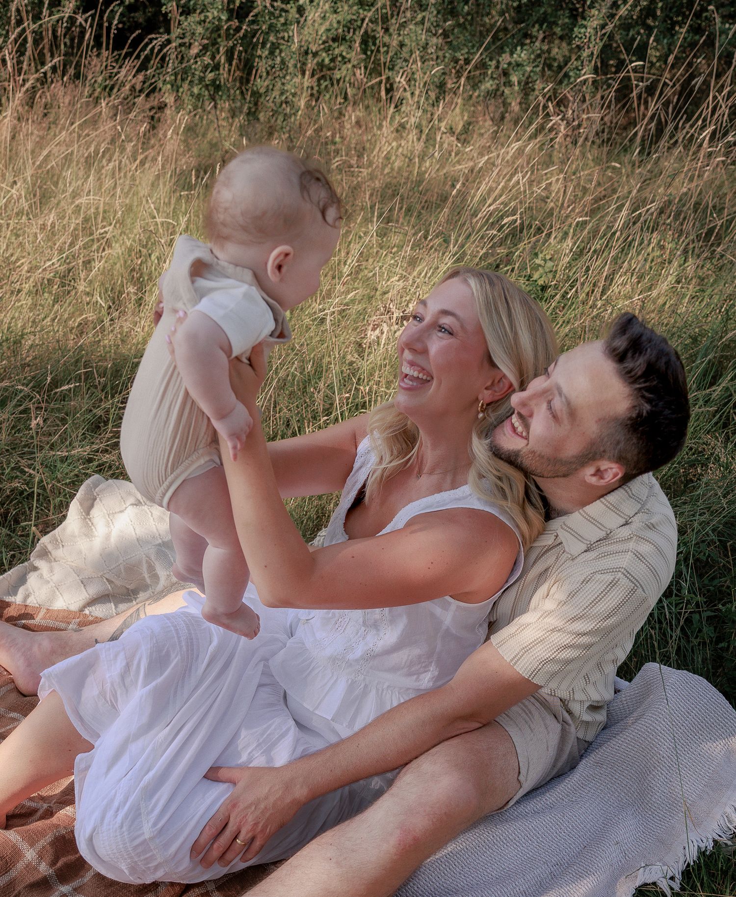 A happy young family sits in a sunny meadow, with parents smiling at their baby in a heartwarming outdoor moment in Stow, Cotswolds.