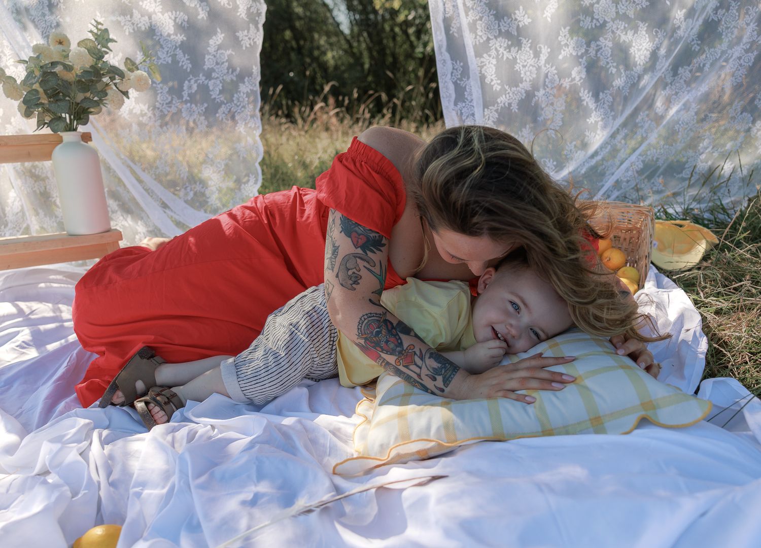 A tattooed mother in a red dress tenderly kisses her toddler lying on a white blanket outdoors in Fairford, Cotswolds.
