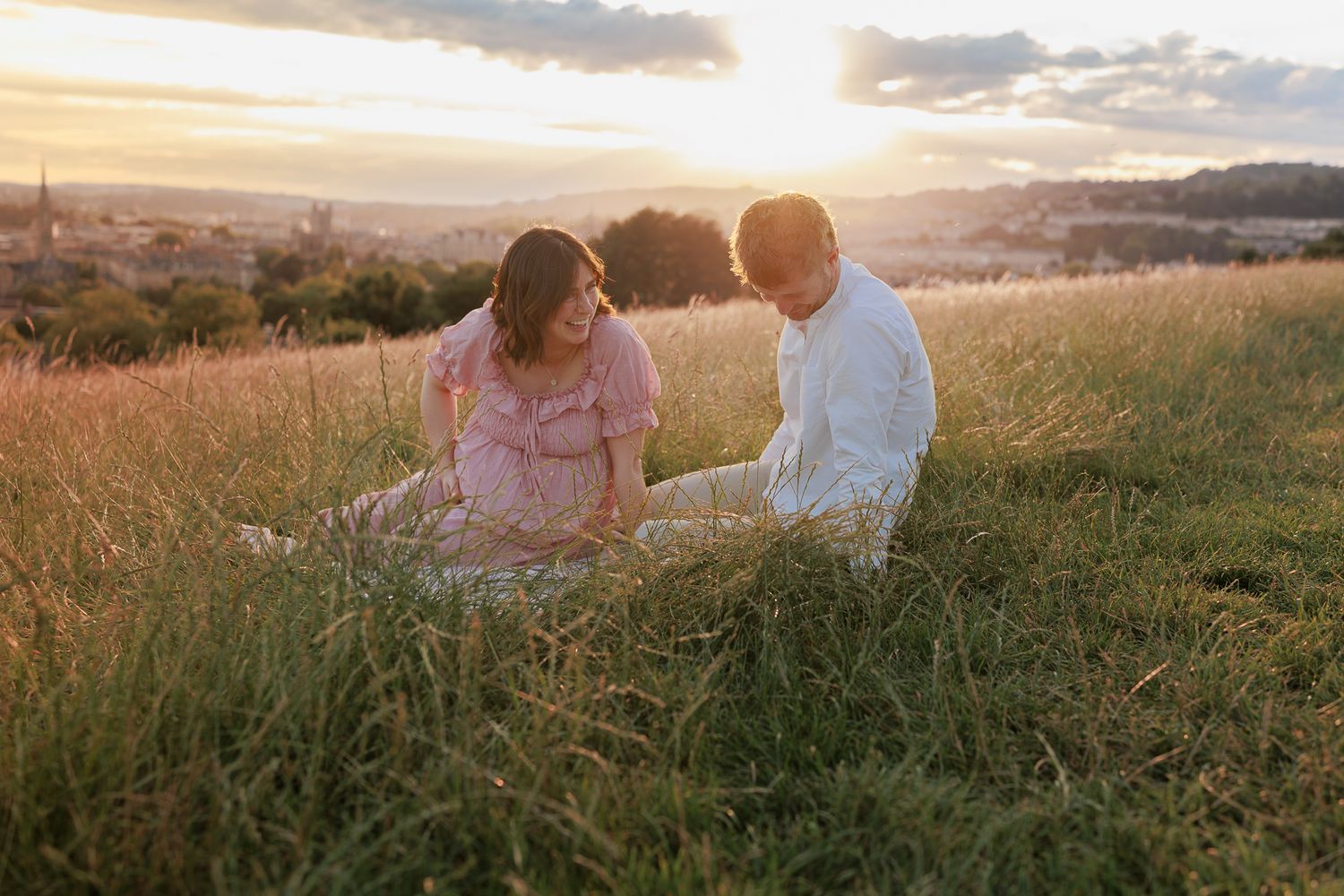 Pregnant couple sitting in golden sunset meadow, woman in pink dress, man in white shirt, Basth city skyline background.
