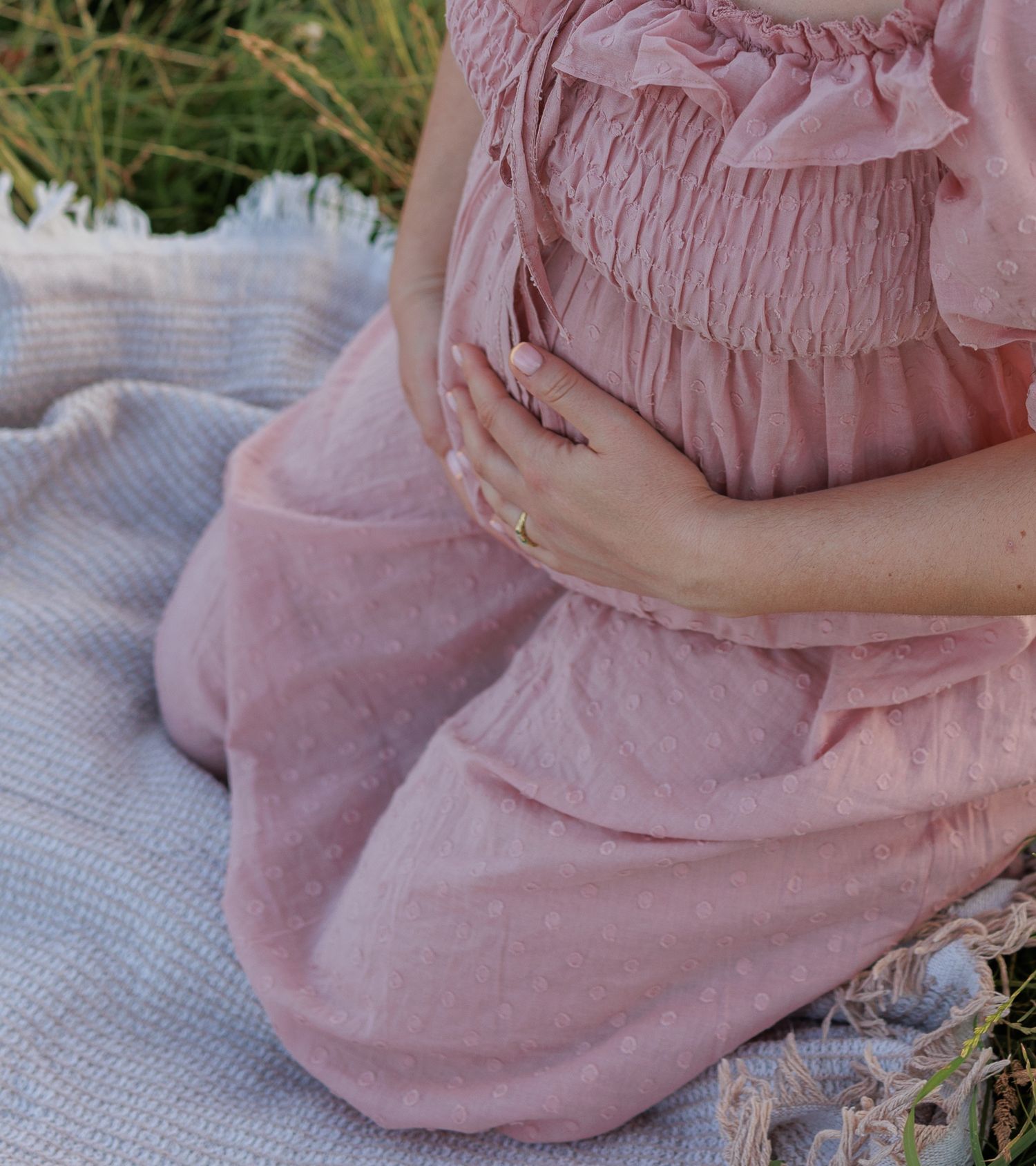 Close-up of pregnant woman in pink smocked dress cradling baby bump on outdoor blanket in Old Windsor, UK.