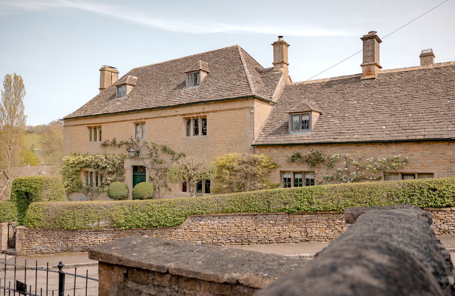 A charming Cotswold stone manor house with climbing plants, dormer windows, and manicured hedges in Upper Slaughter.