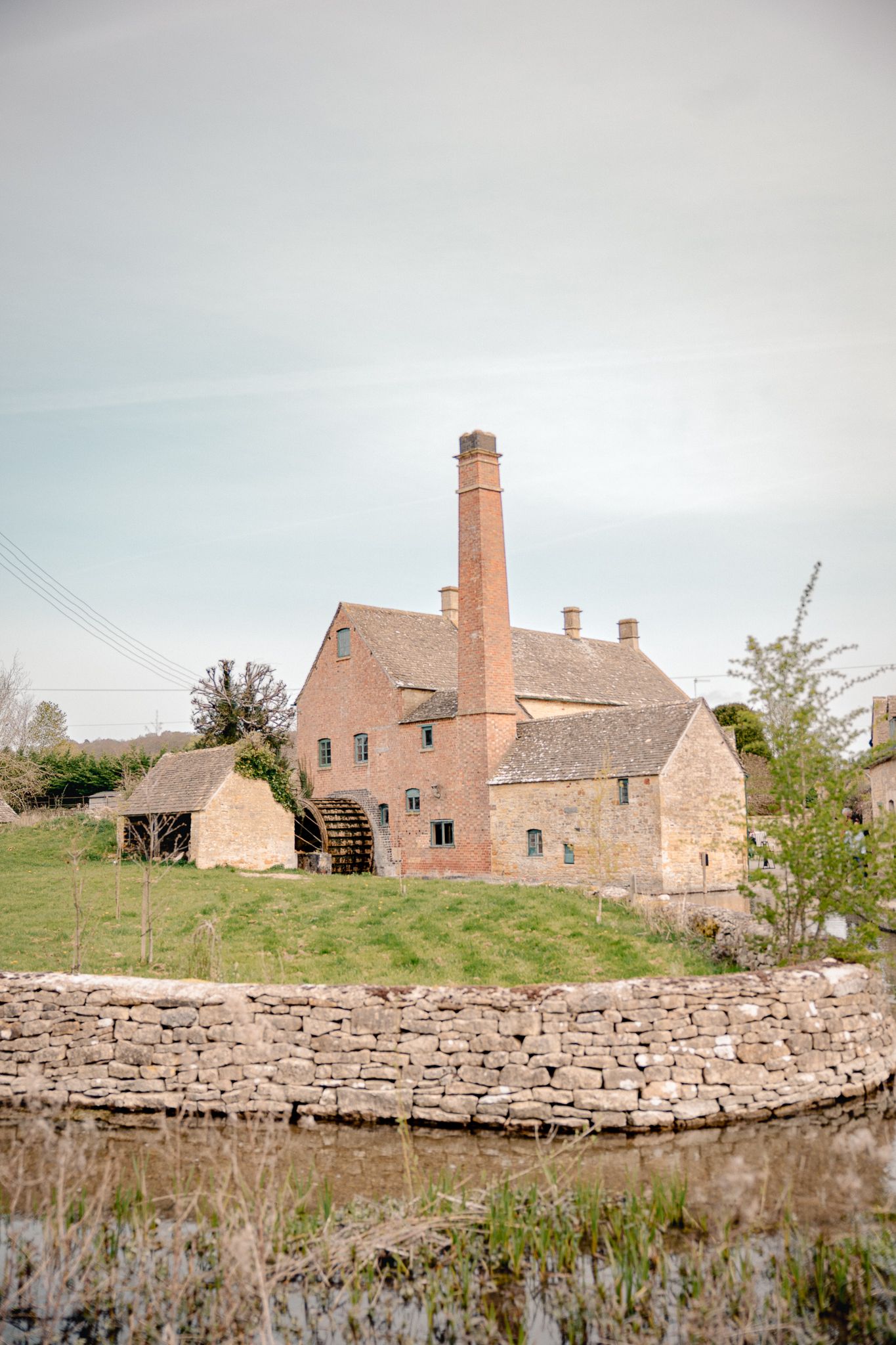 Historic brick watermill with tall chimney beside a stone wall and millpond in the English Cotswolds, Lower Slaughter Museum.