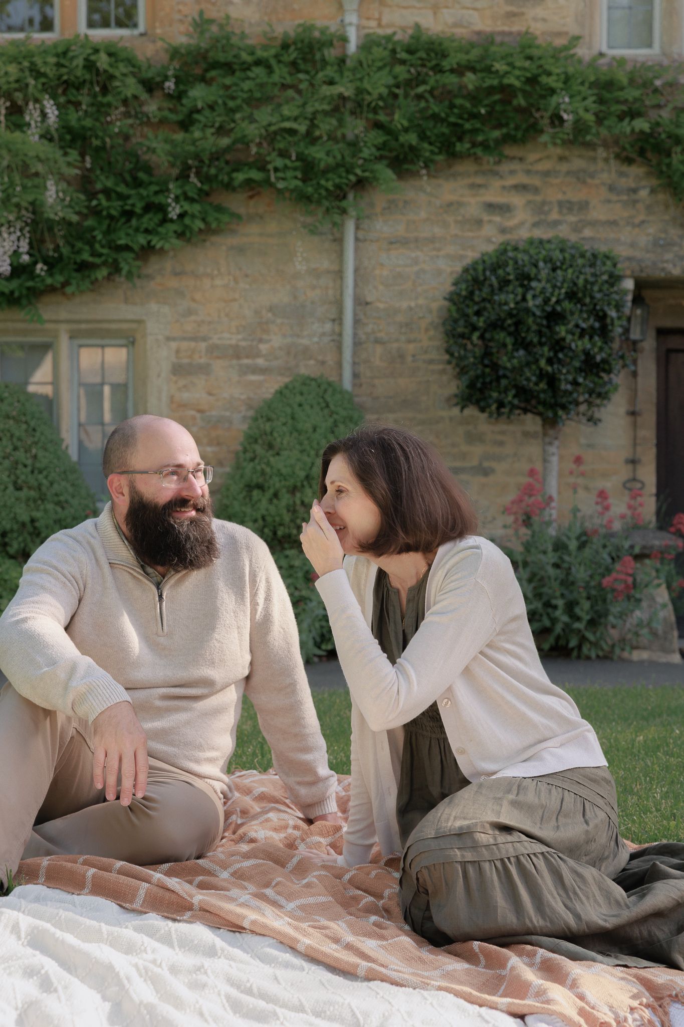 Couple sharing an intimate conversation on a blanket in a lush garden outside a stone cottage.