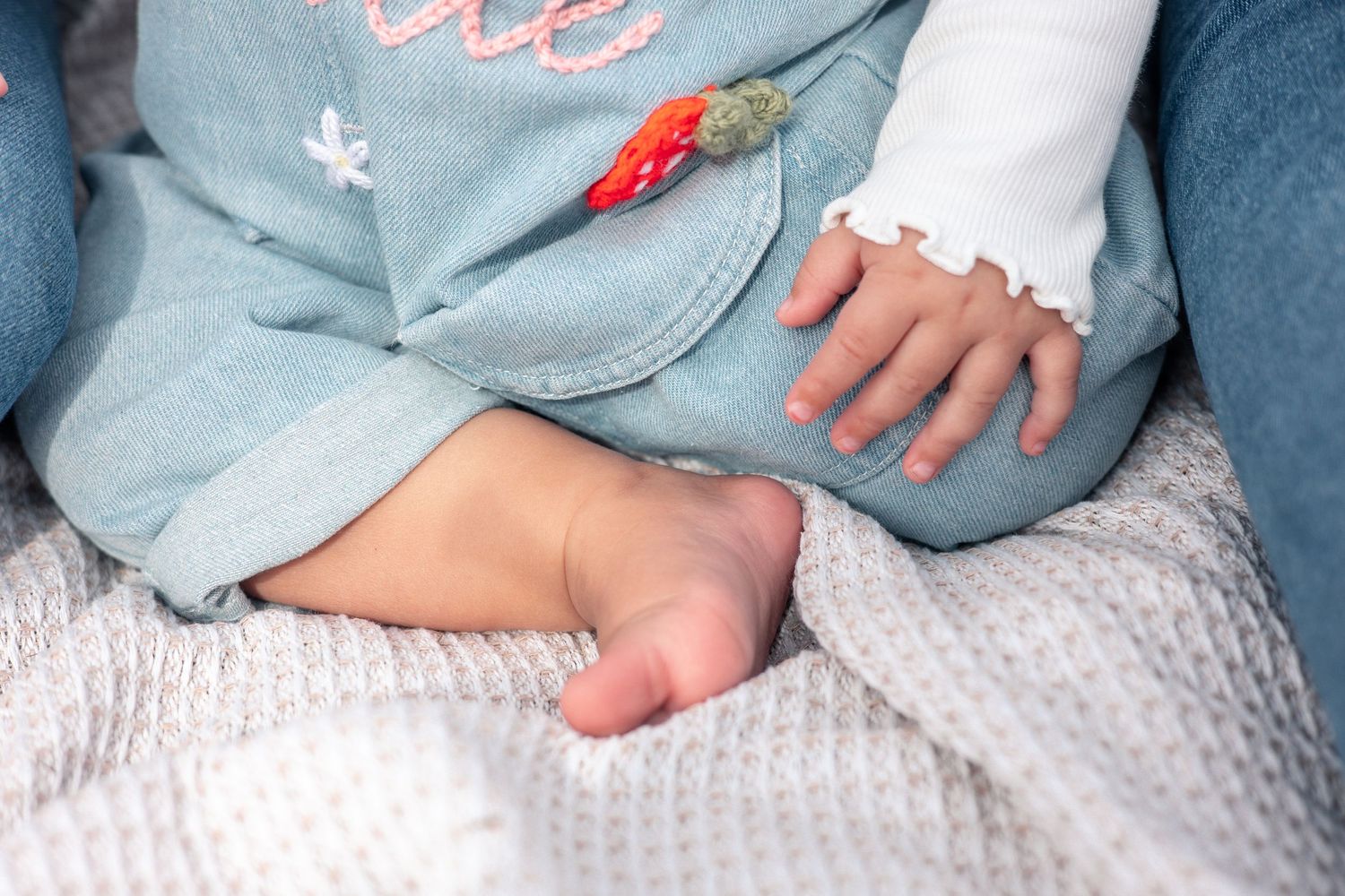 Close-up of a baby's bare foot and hand resting on a soft white blanket, wearing a light blue outfit.