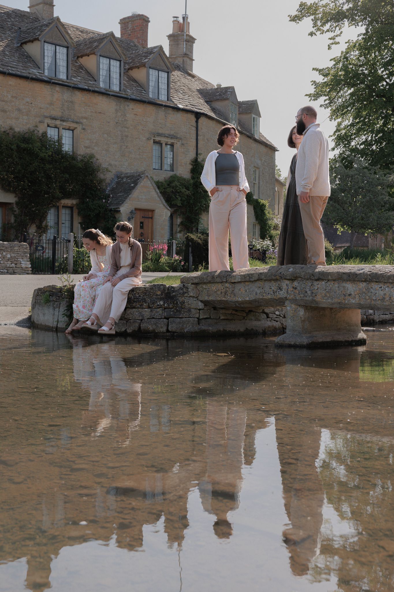 Four people gather on a stone bridge over a reflective pond beside a charming Cotswolds village.