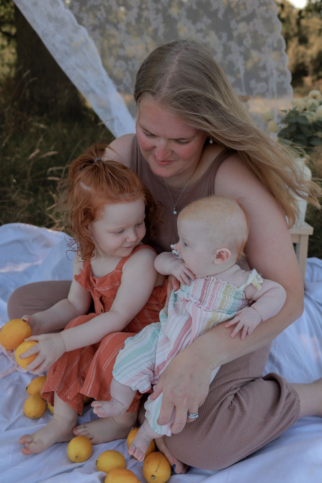 A mother smiles at her two young children, a toddler in orange and a baby, sitting outdoors with lemons in Henley-on-Thames, Oxfordshire.