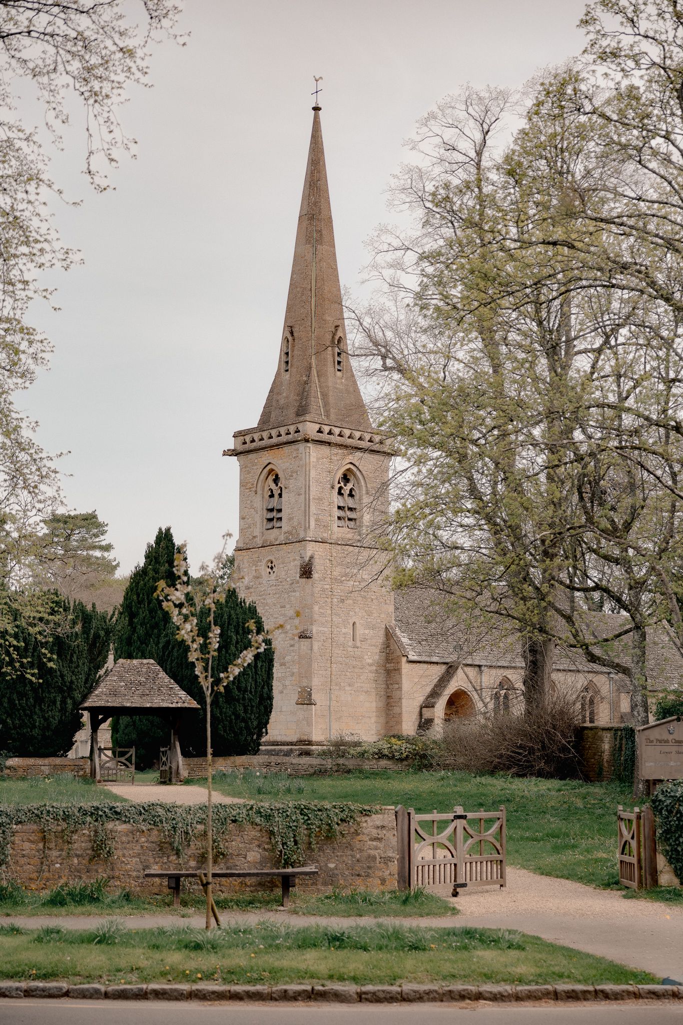 Historic stone church with pointed spire surrounded by trees and wooden gate in an English countryside village in Snowshill.
