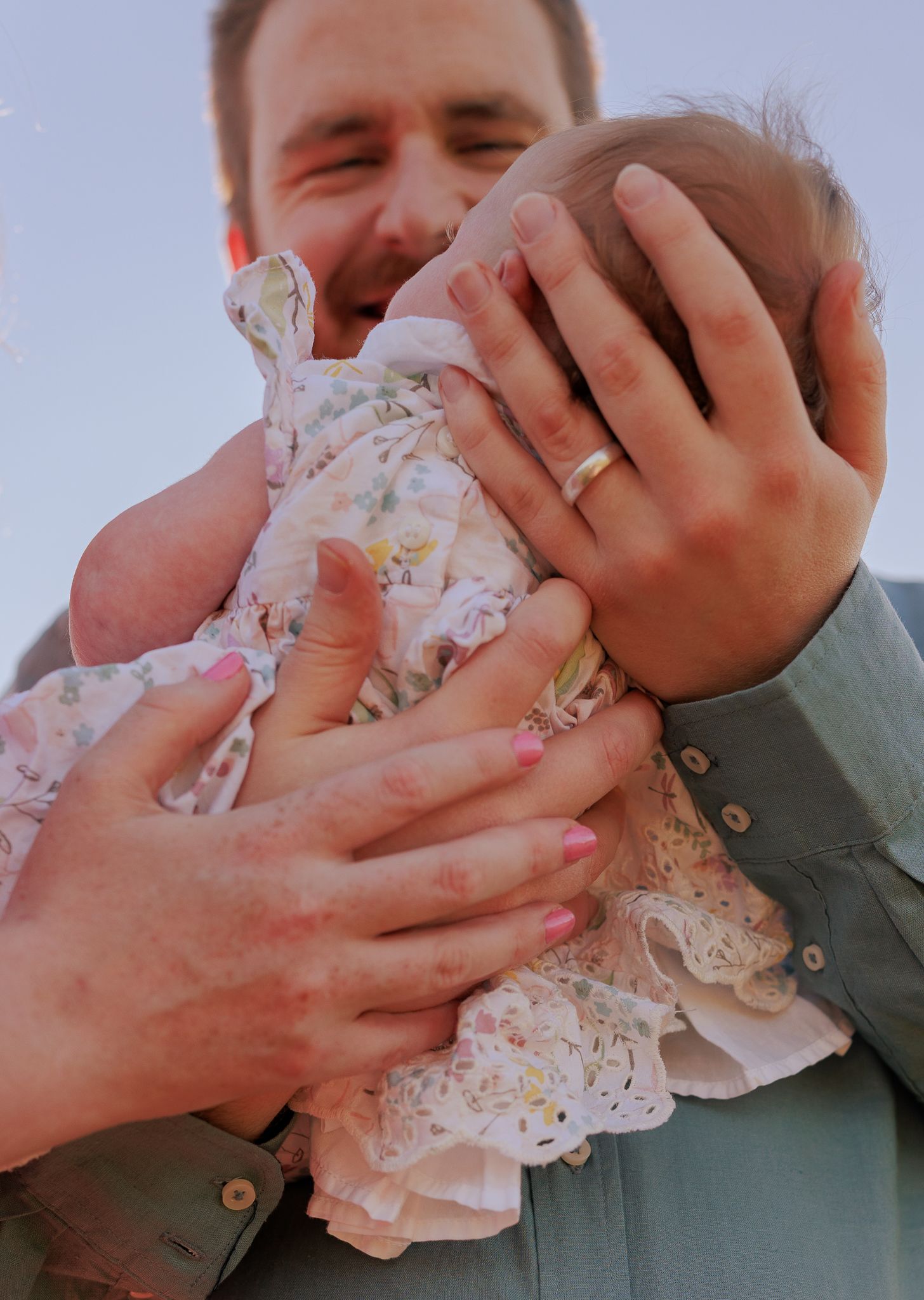 A father holding his newborn baby in a floral outfit, smiling joyfully, wearing a wedding ring outdoors in New Milton.