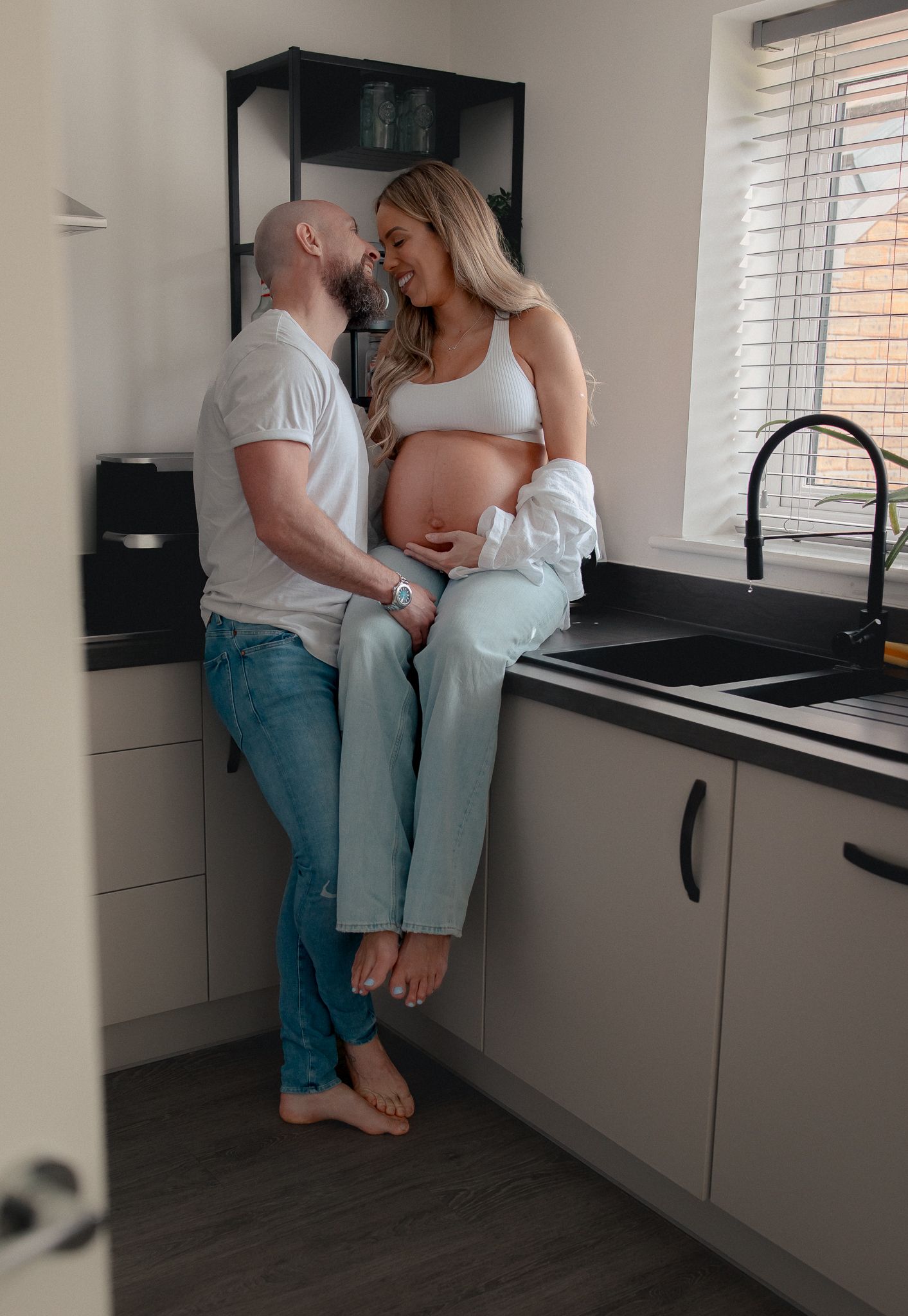 Couple sharing a tender moment in kitchen, pregnant woman sitting on counter cradling baby bump, smiling at partner in Coln St Aldwyns.