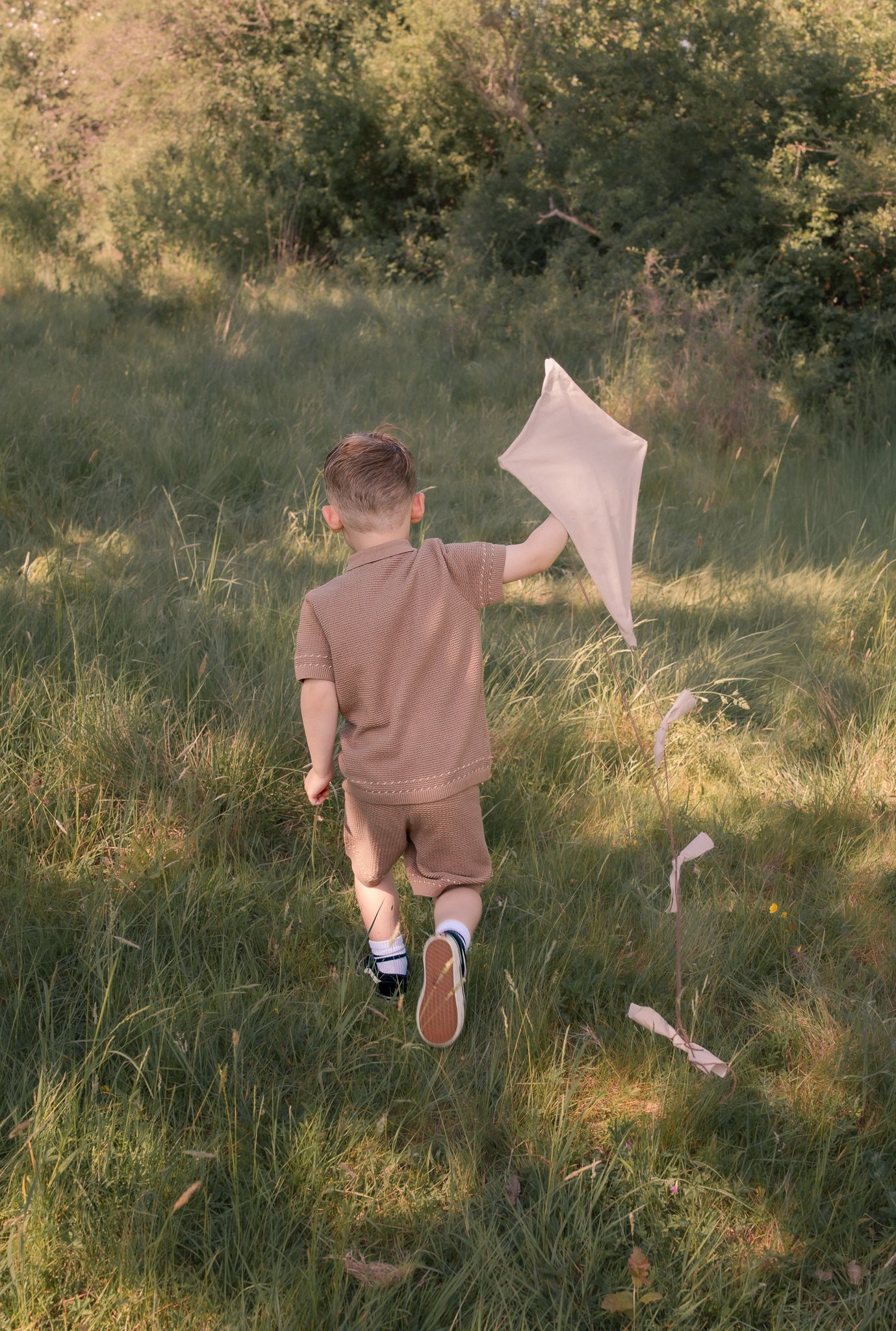 A young boy in a tan outfit runs through a green meadow holding a butterfly net on a sunny day.