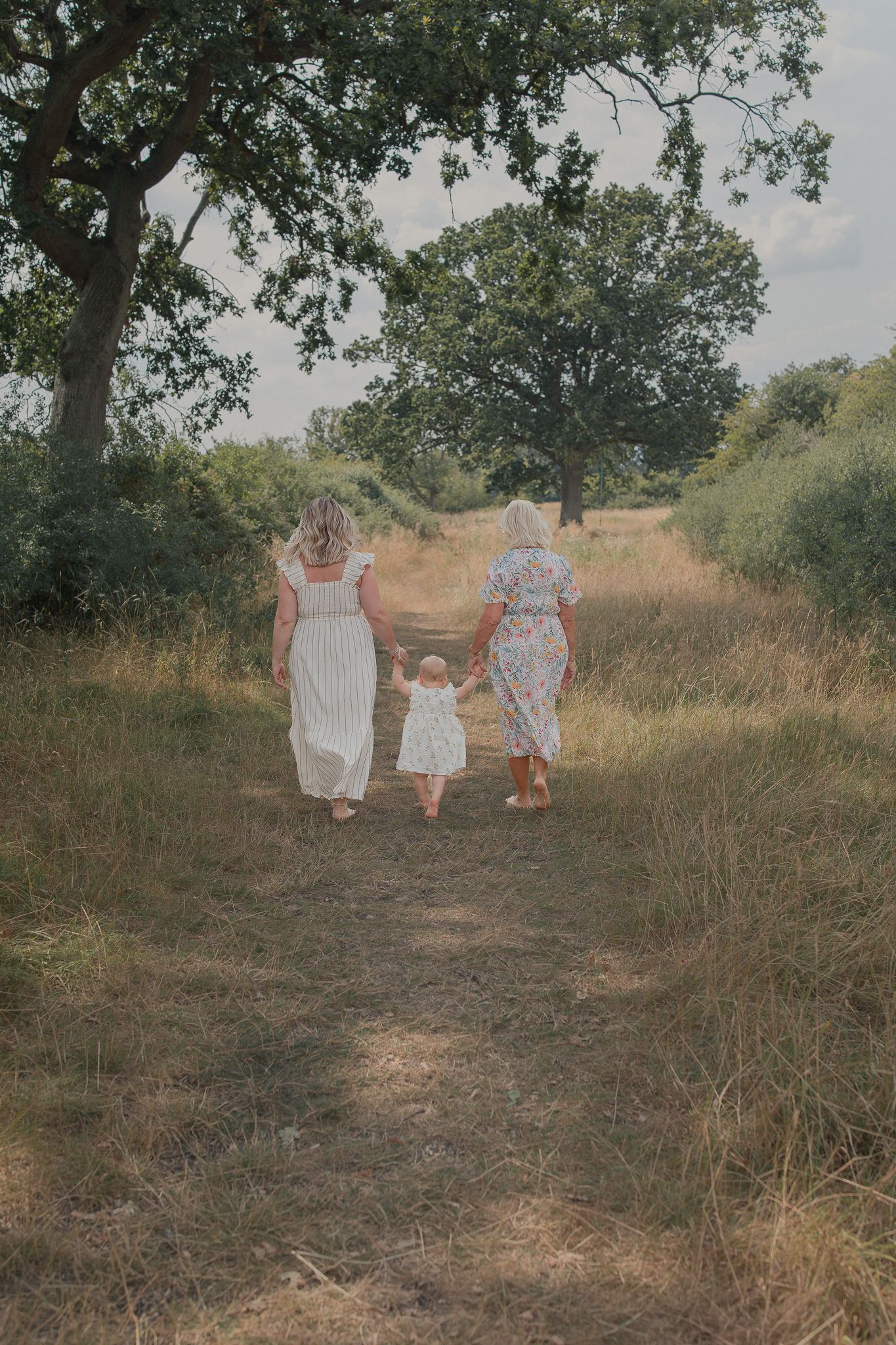 Two women in floral dresses walk hand-in-hand with a toddler along a sunlit countryside path surrounded by lush green trees.
