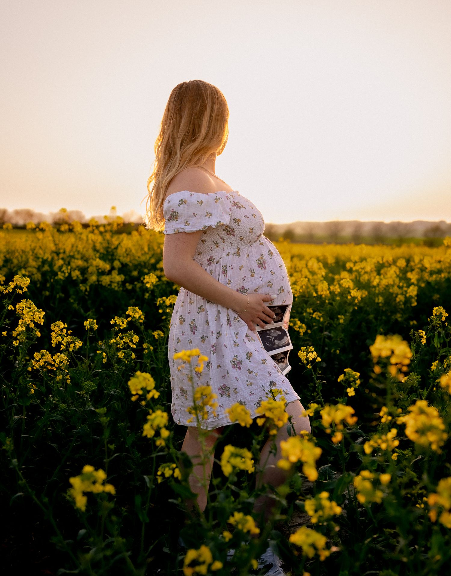 Pregnant woman in floral dress holding ultrasound photo in golden yellow flower field at sunset in Witney.