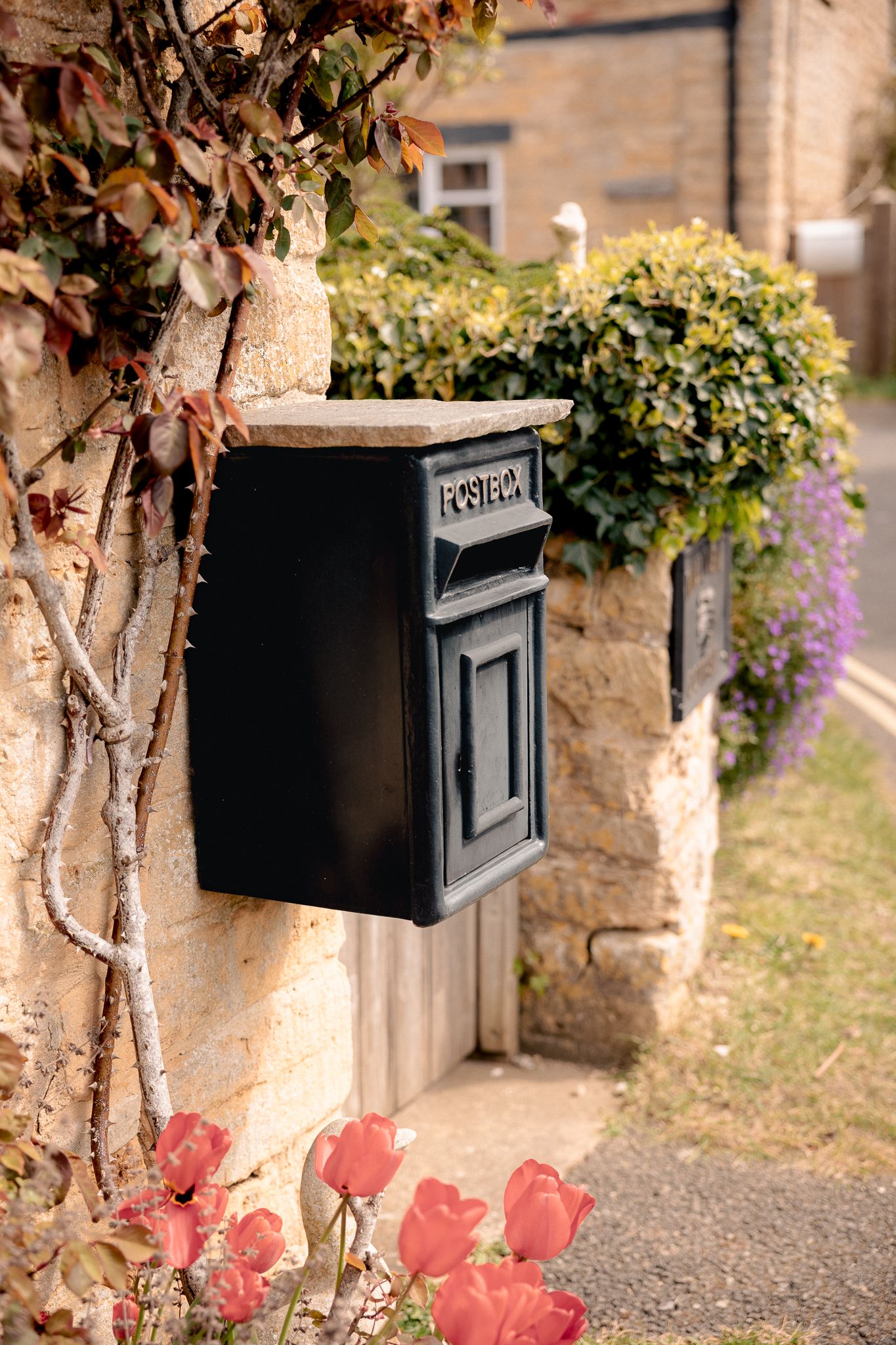 Black cast iron postbox mounted on a stone wall with red roses and ivy in an English cottage garden in Lower Slaughter, Cotswolds, UK.