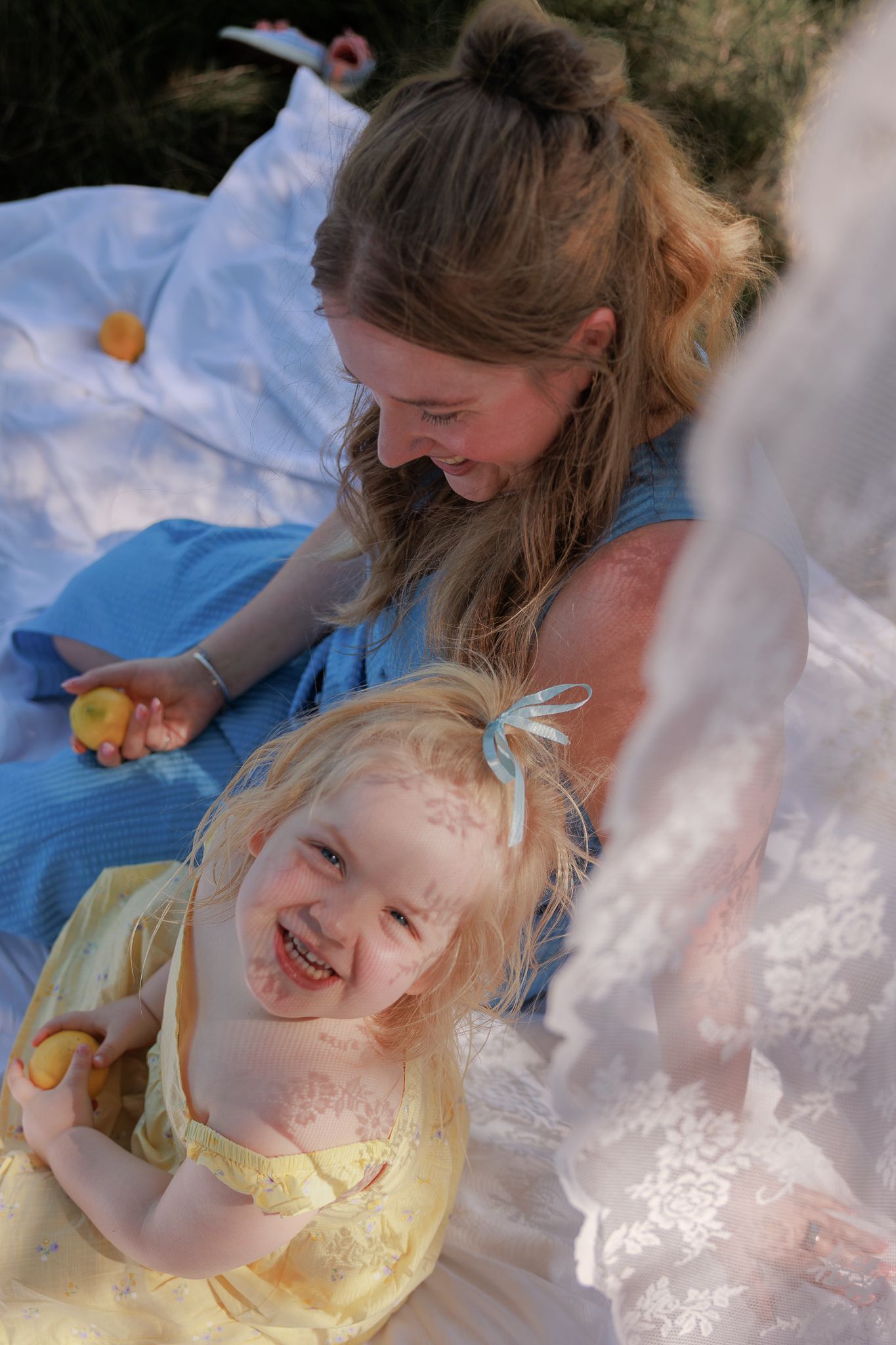 Two young girls laughing and playing outdoors on a white blanket, wearing blue and yellow dresses at a picnic in Bibury, Cotswolds.