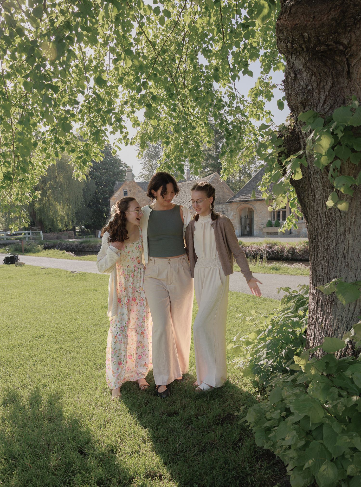 Three young women walk together under a leafy tree in a sunlit green garden near a stone cottage.