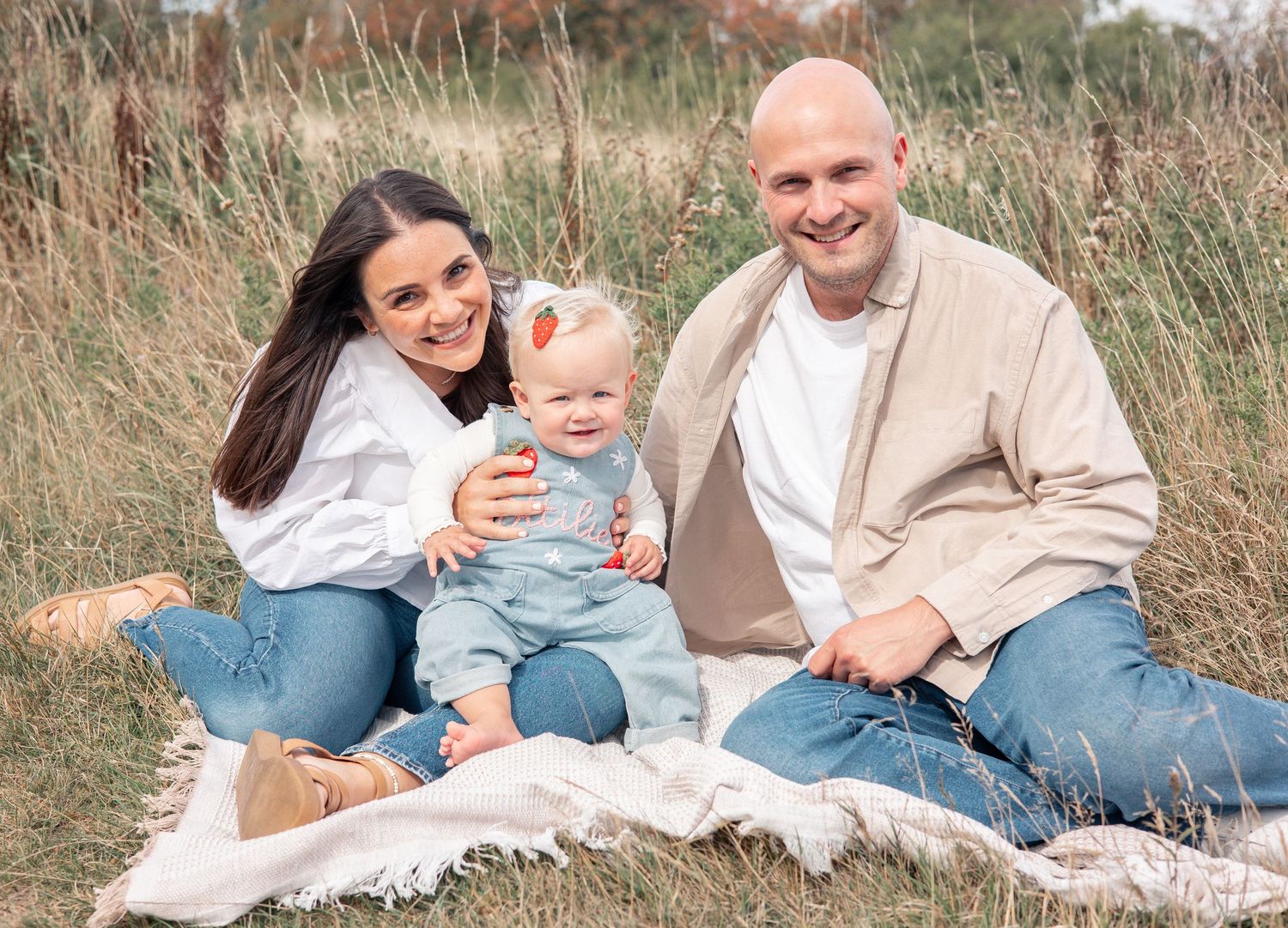 Happy family of three sitting together outdoors in a field with tall grass, wearing coordinating neutral outfits.