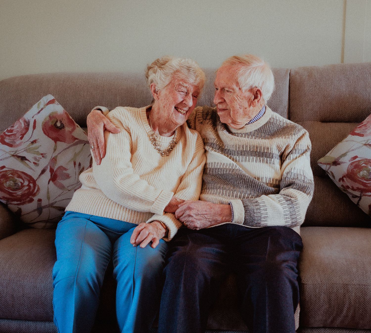Elderly couple sitting on a sofa, smiling and embracing each other warmly in Moreton-in-Marsh.