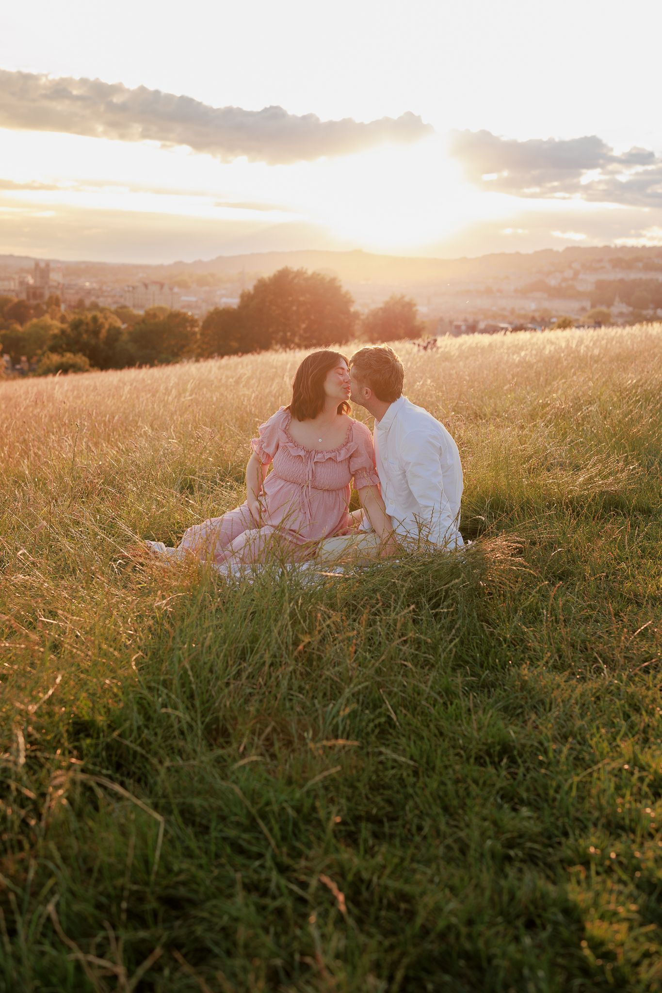 Couple sitting in golden sunset meadow grass, for a romantic maternity Maternity Photoshoot in Clevedon.